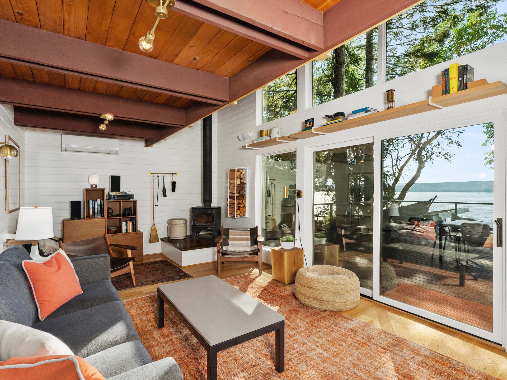 A cozy living room with large windows revealing a serene lake view. The room features a gray sofa with orange cushions, a wooden coffee table, a wood-burning stove, and rustic decor. Sliding glass doors lead to a deck with outdoor seating.