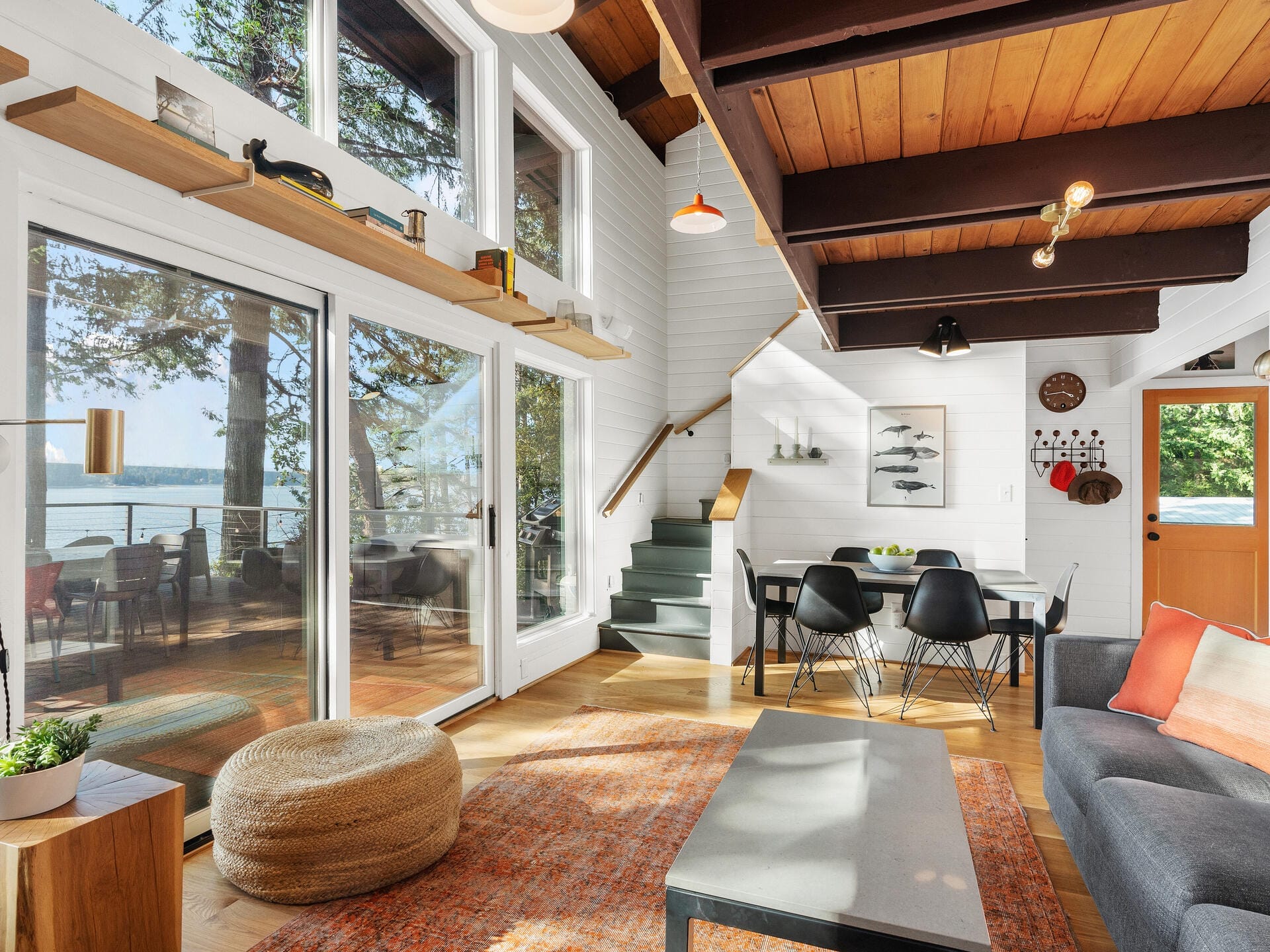 A bright and airy living room with large windows and a sliding door opening to a patio. It features a wooden ceiling with beams, modern furniture, a pouf, and a dining area. The space is decorated with plants and artwork, and offers views of the outdoors.