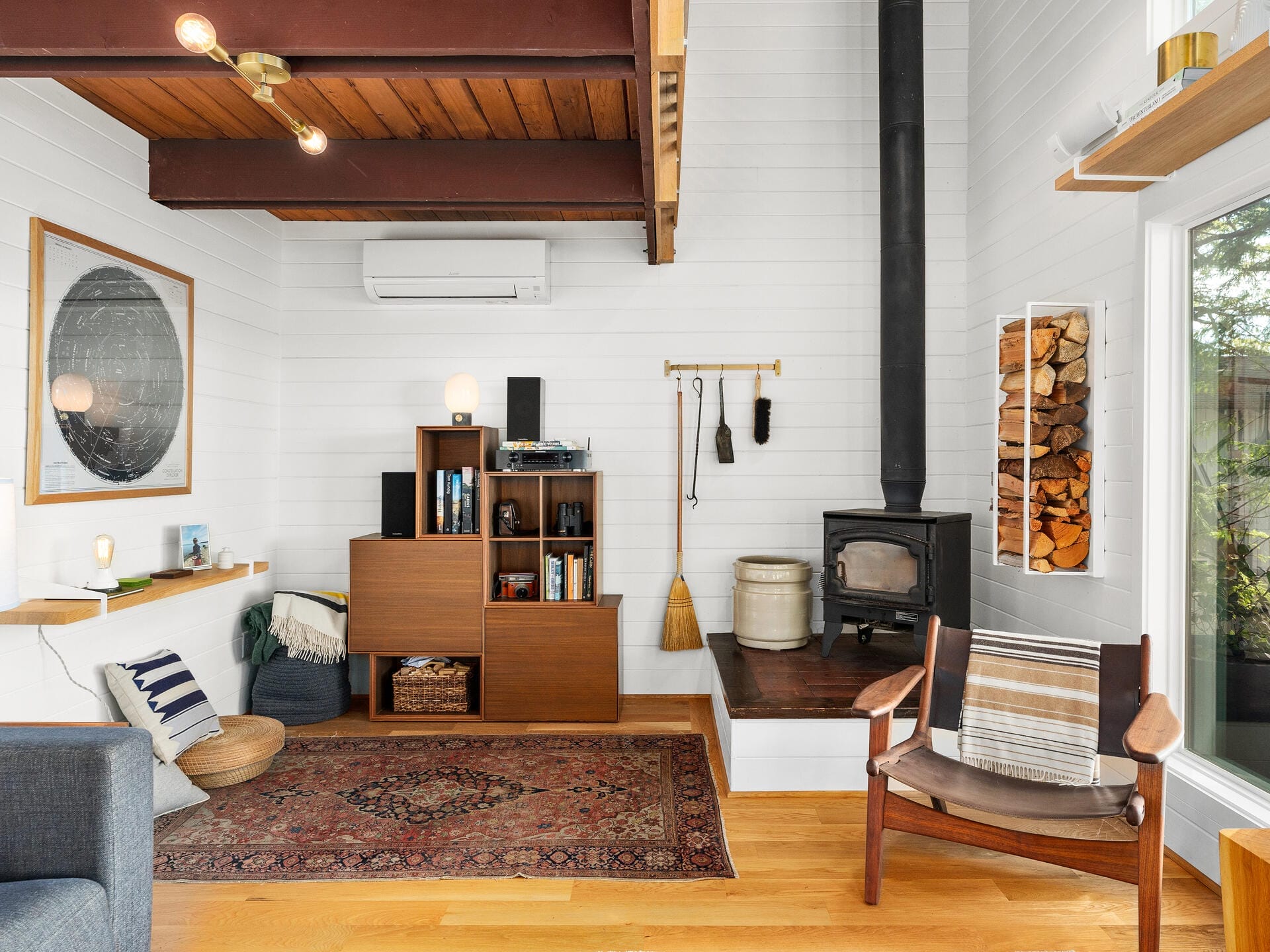 Cozy living room with a wood-burning stove, bookshelf, and a large rug. A rocking chair is beside a stack of firewood. White walls and wooden ceiling beams create a warm atmosphere. A large window lets in natural light.