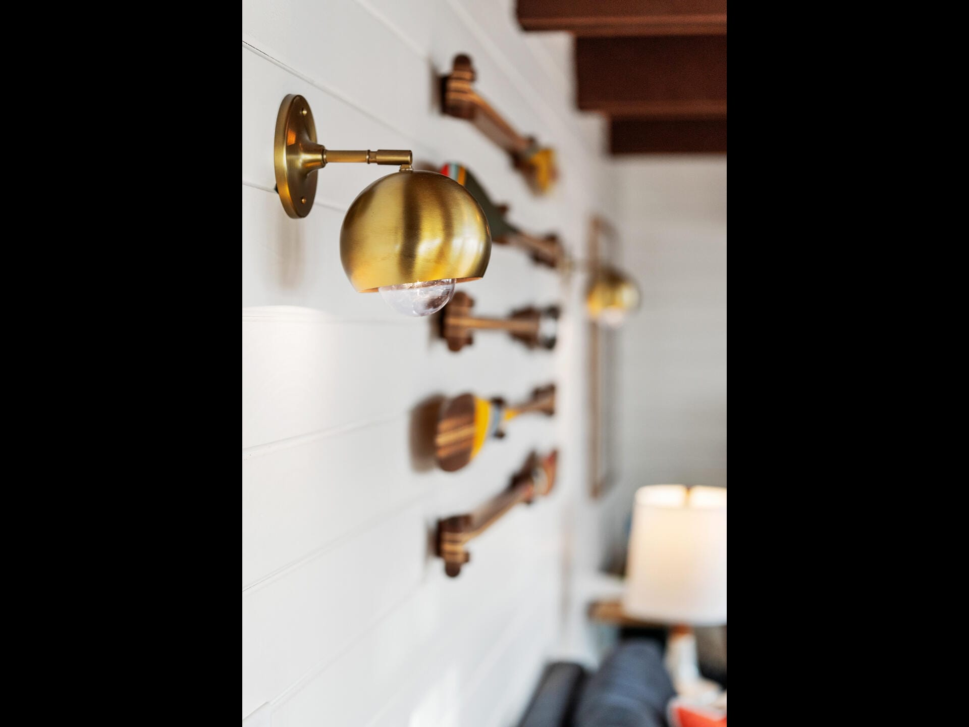 A close-up of a golden wall-mounted lamp on a white shiplap wall. In the background, several oars are displayed decoratively. A soft-focus living room setting is partially visible, showing a lamp and part of a couch.