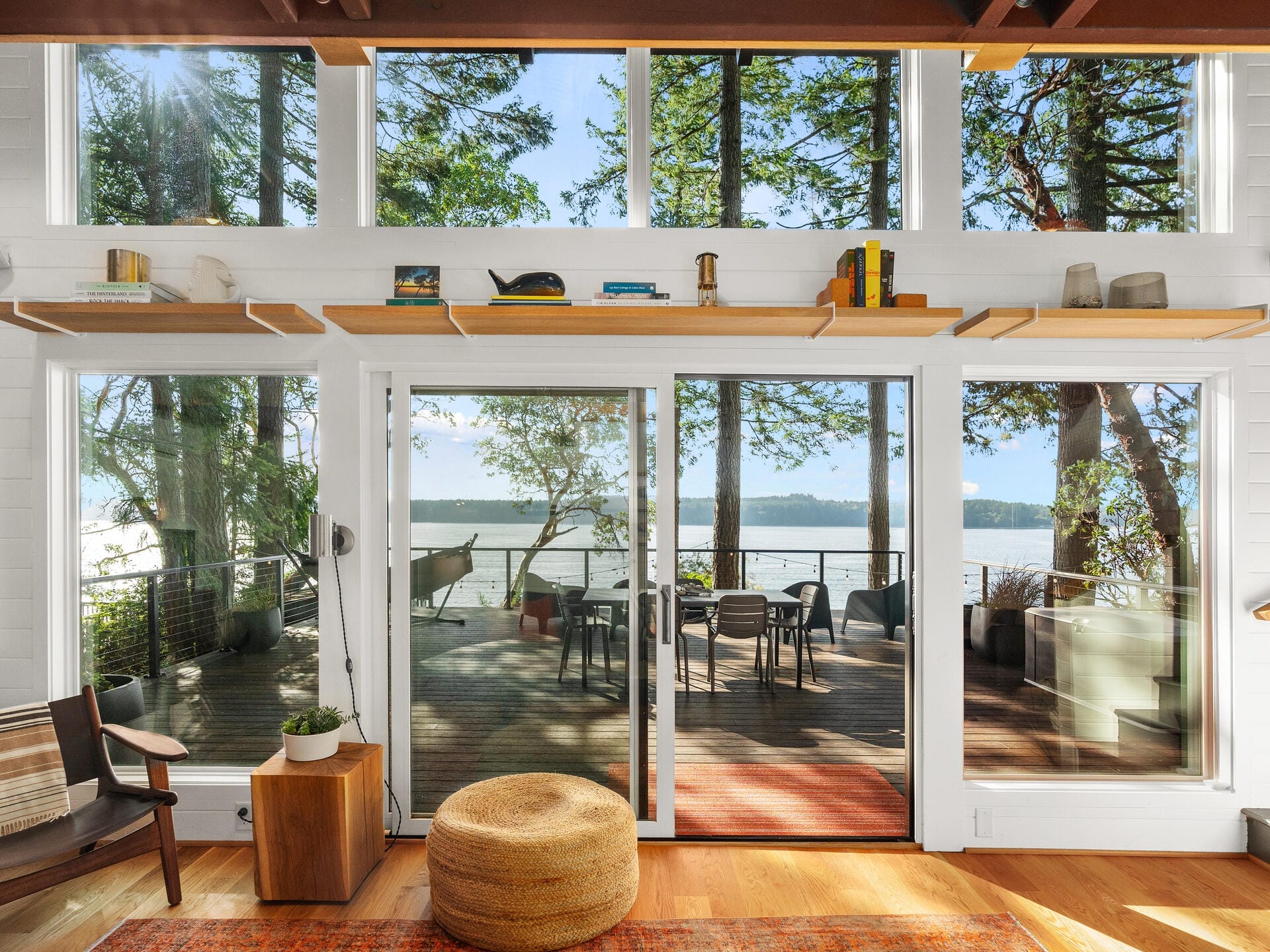 A bright and airy room with large windows overlooking a lakeside deck. The room has light wooden flooring, a woven pouf, and minimalist decor. The deck features outdoor seating and a hammock, surrounded by trees and a view of the water.