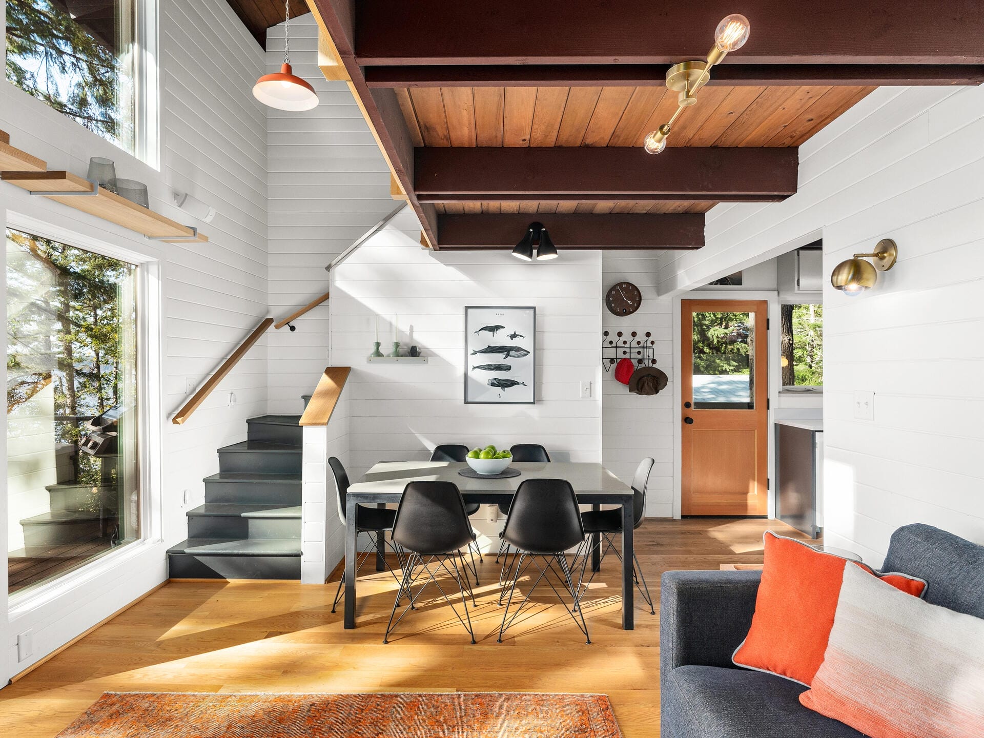 A modern, open-plan living and dining area with wooden floors and white walls. It features a black dining table with black chairs, a gray sofa with colorful cushions, and a staircase. Natural light streams in through large windows.