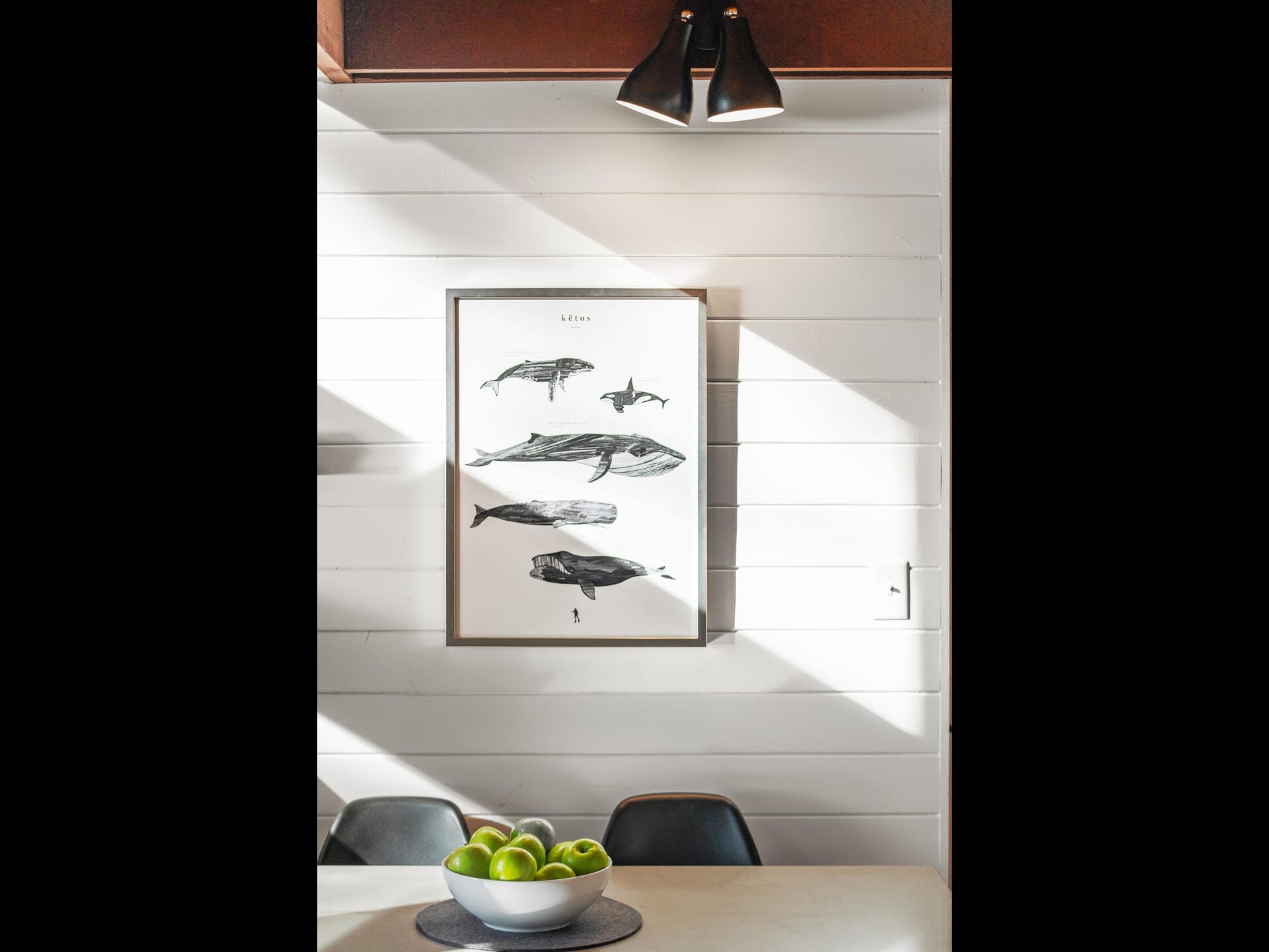 A framed illustration of various whales hangs on a white shiplap wall. Below, a light gray table holds a bowl of green apples, with two black chairs partly visible. Soft sunlight filters through, creating a warm, inviting atmosphere.