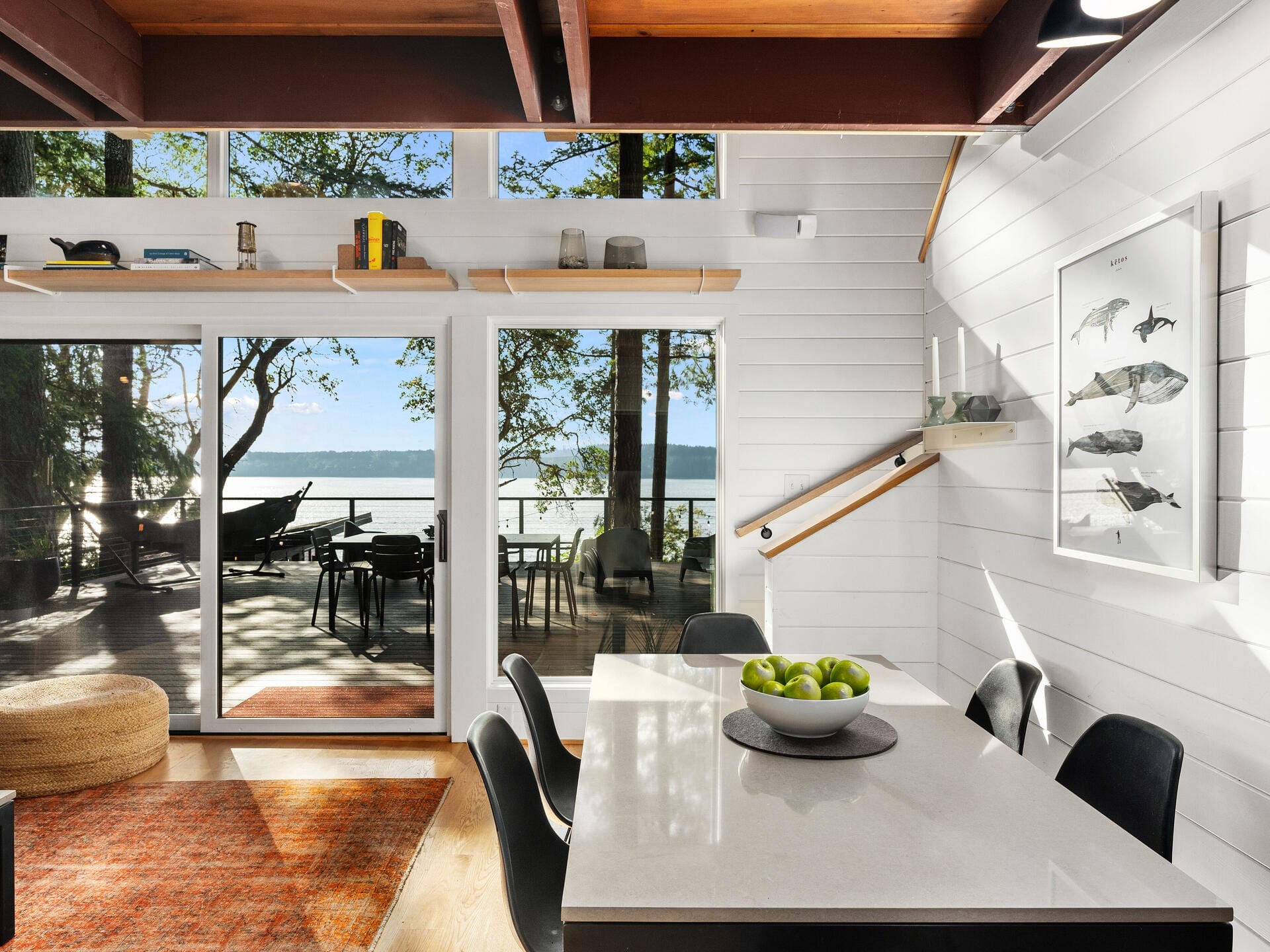 A modern, sunlit dining area with a white table and black chairs. A bowl of green apples sits on the table. Glass doors open to a deck with lounge chairs, overlooking a serene lake. Wooden beams contrast with white walls adorned with a fish-themed artwork.