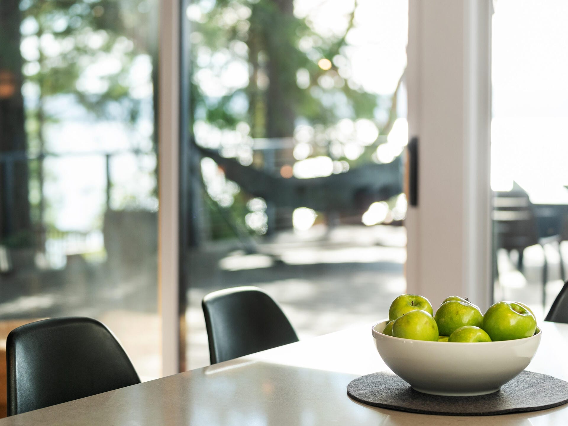 A sunlit kitchen area featuring a bright tabletop with a white bowl filled with green apples. In the background, glass doors open to a patio with a blurred view of a hammock, trees, and outdoor furniture.