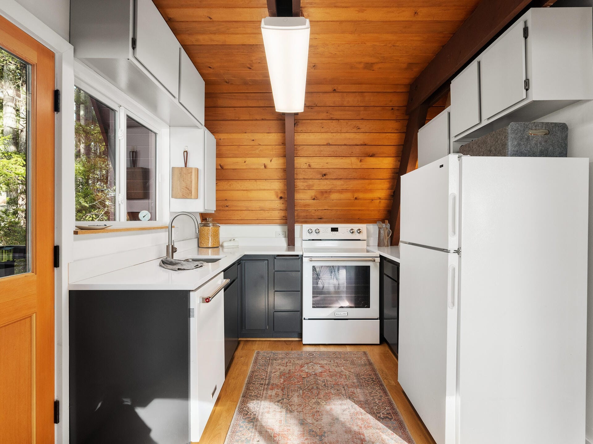 A cozy kitchen with white cabinets, dark lower cabinets, and a wooden slanted ceiling. It features a white refrigerator, stove, and dishwasher, with a large window on the left. A patterned rug covers the light wooden floor.