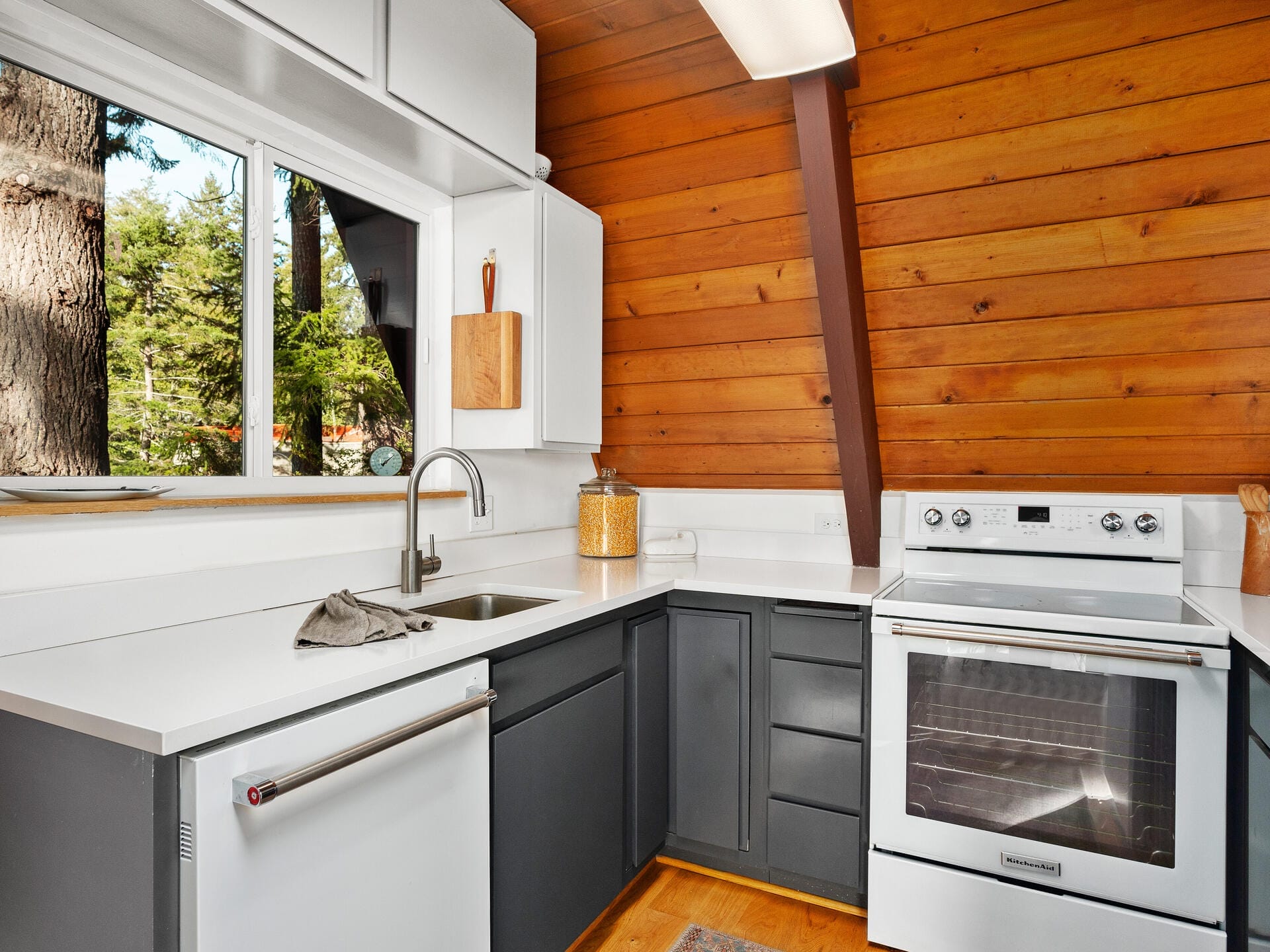 A modern kitchen with white countertops, dark gray lower cabinets, and wood-paneled walls. It features a stainless steel sink, stove, and dishwasher. Large window with a view of trees. A cutting board and jar are on the counter.