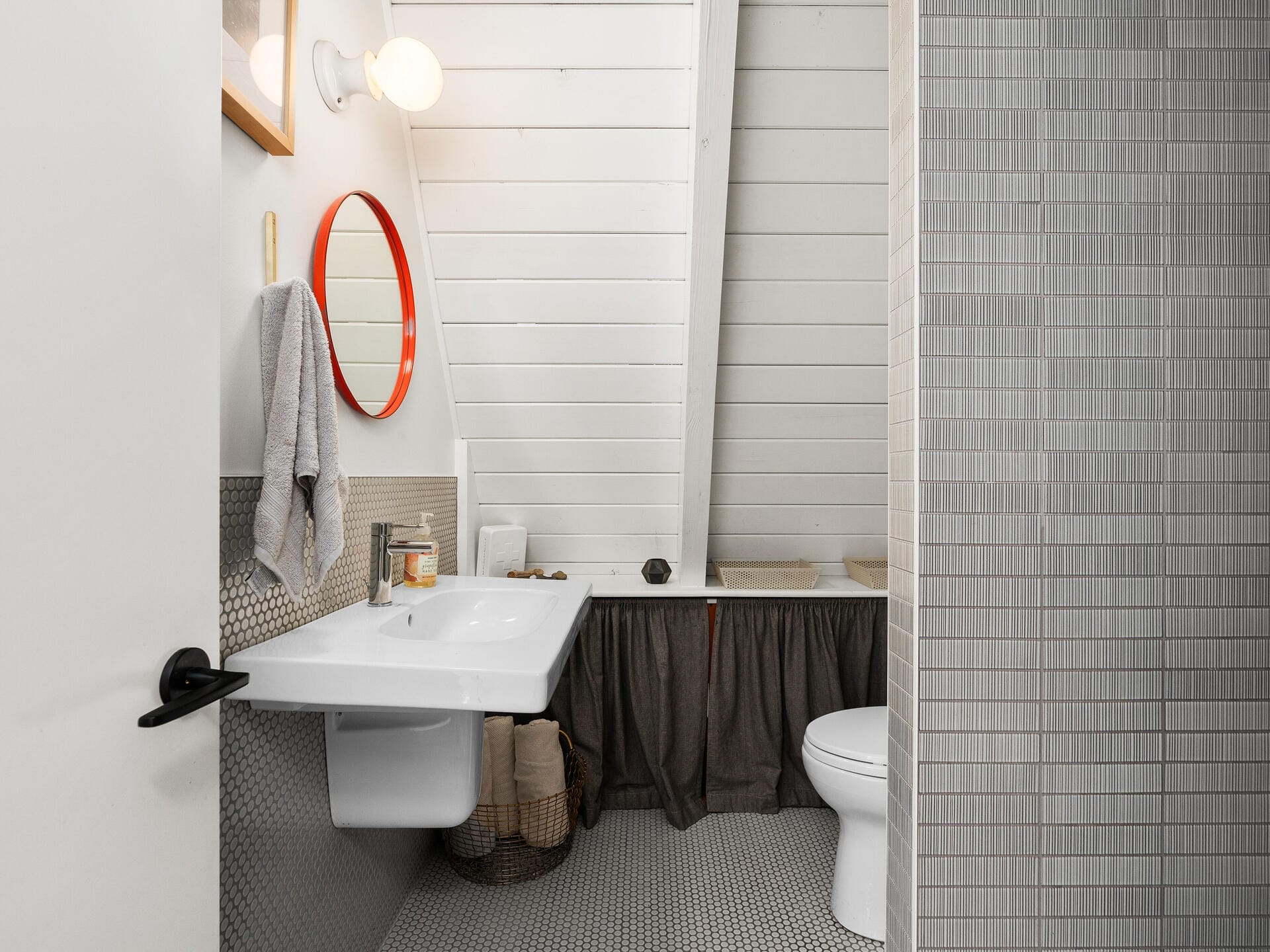 A modern bathroom with a white sloped ceiling. It features a wall-mounted sink, a red circular mirror, a gray towel, a toilet, wicker baskets, and a shower behind a gray tiled partition. The floor has small, round gray tiles.