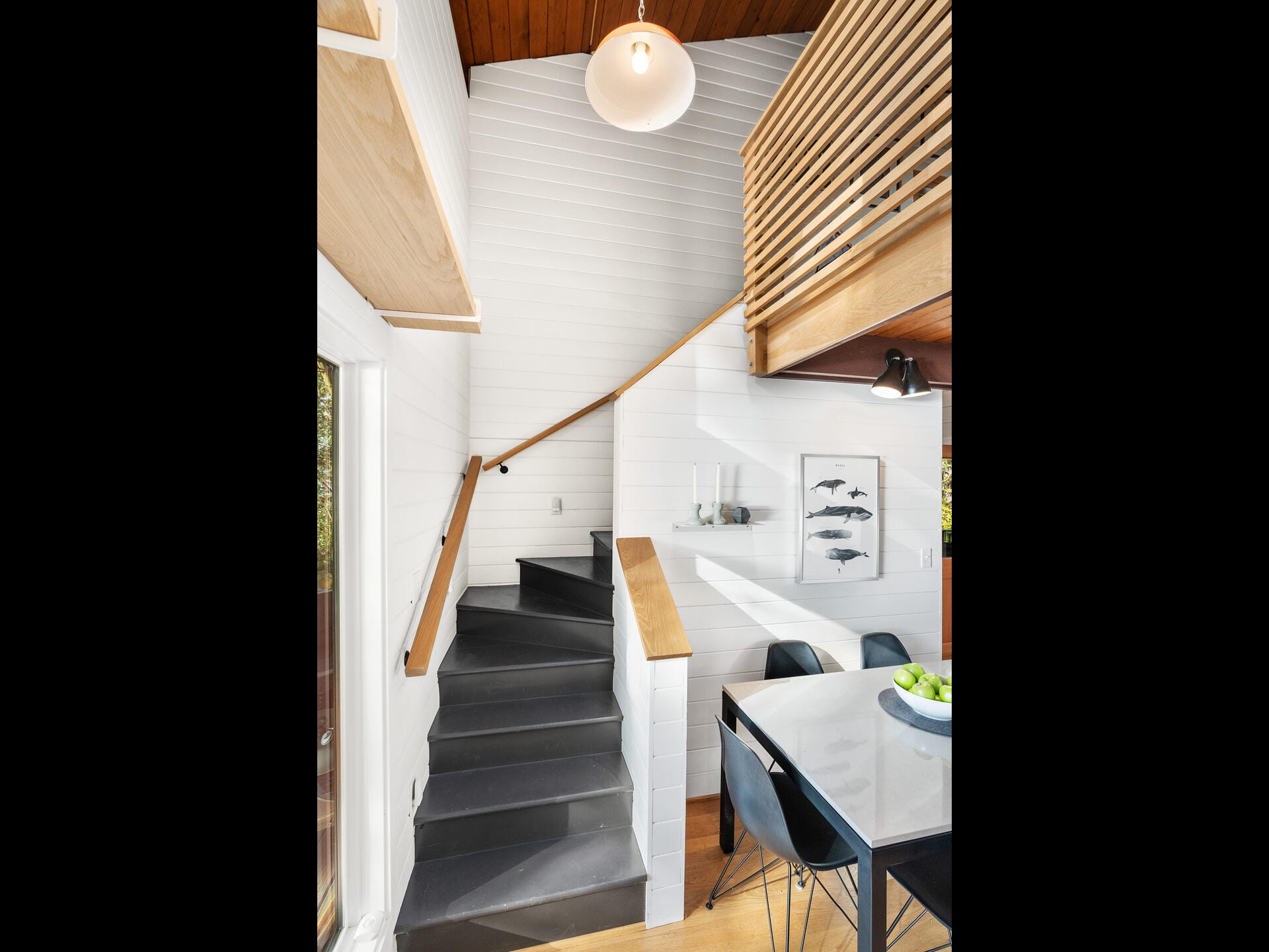 A modern interior features a white-walled room with a wooden ceiling. A winding staircase with a wooden handrail leads up. Below, a dining table with black chairs and a centerpiece is visible. A pendant light hangs above. Decor includes framed fish prints.