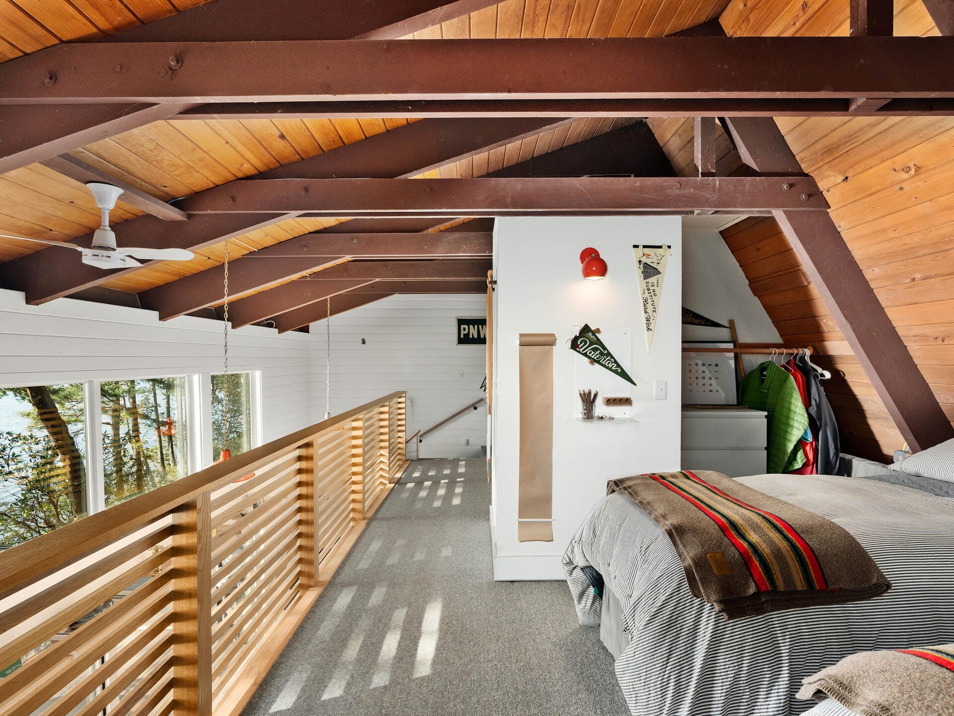 A cozy loft bedroom with a slanted wooden ceiling, skylights, and exposed beams. The room features a bed with a striped blanket, a wall with a pennant, and a view of trees through the railing. Carpeted flooring and natural light enhance the warmth.