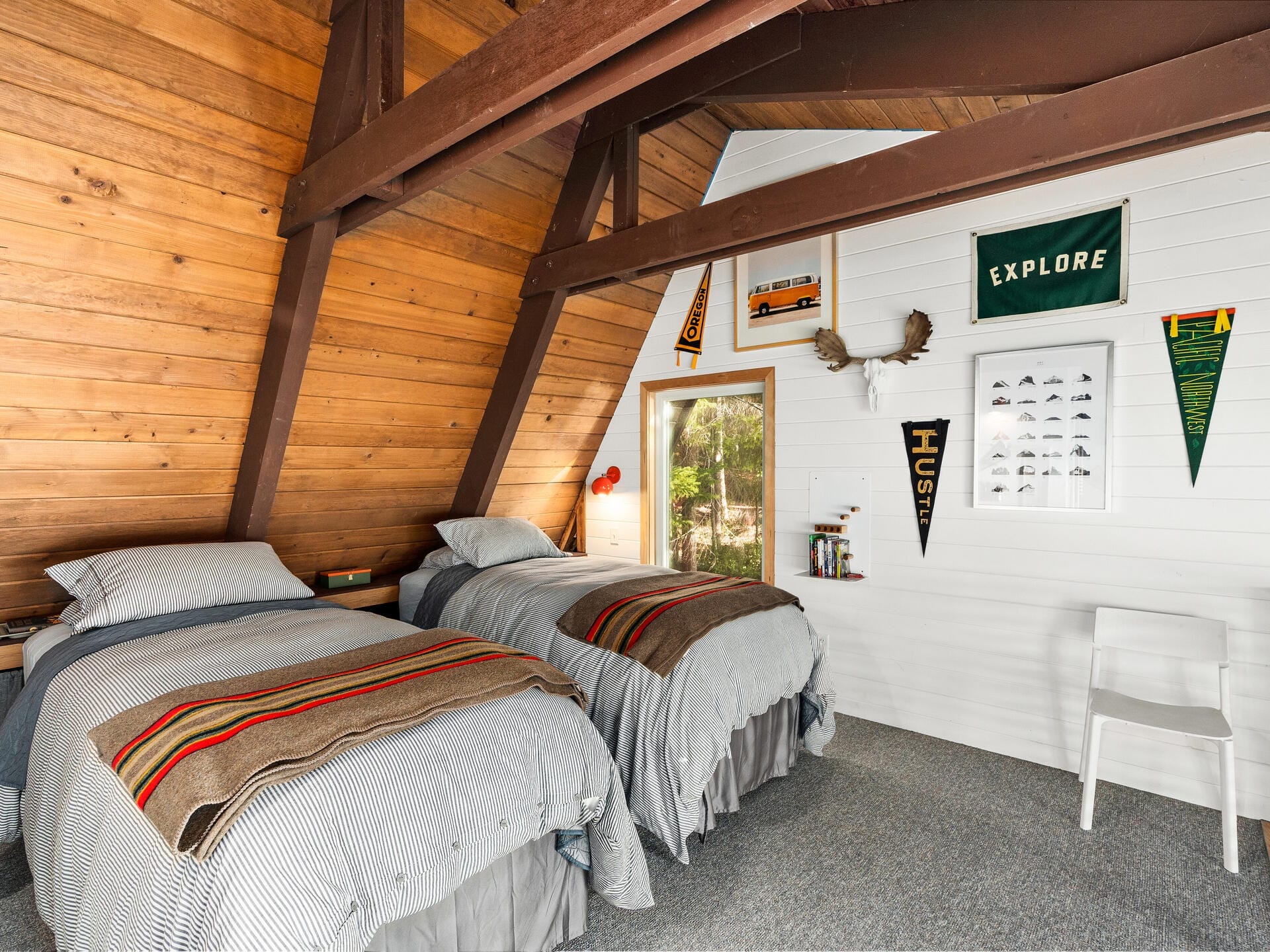 A cozy attic bedroom with two twin beds, each covered with striped blankets. The wooden walls are adorned with banners, a shelf with miniatures, a pair of antlers, and a window offering a view of trees outside. A white chair is placed near the wall.