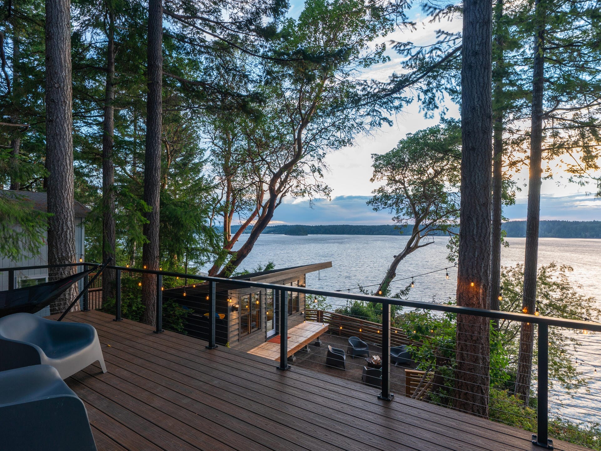 A wooden deck overlooking a serene body of water is surrounded by tall trees. A modern cabin with large glass windows is visible below. The sky is partly cloudy, and the scene is bathed in soft evening light.