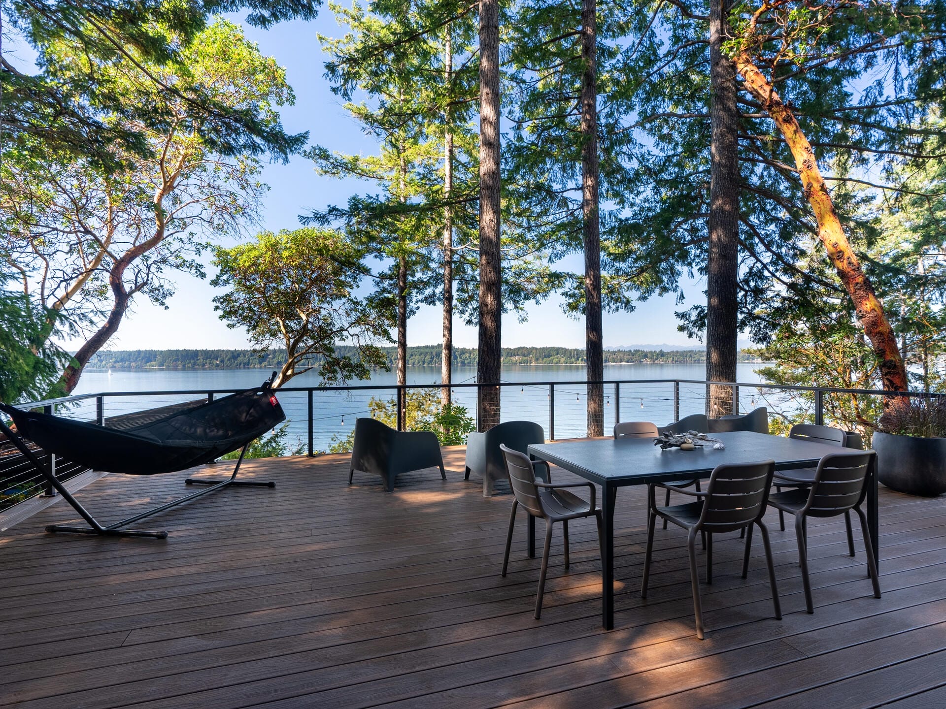 A wooden deck overlooking a serene body of water, surrounded by trees. The deck features a table with chairs, a hammock, and lounge chairs, creating a peaceful outdoor setting with a clear blue sky overhead.