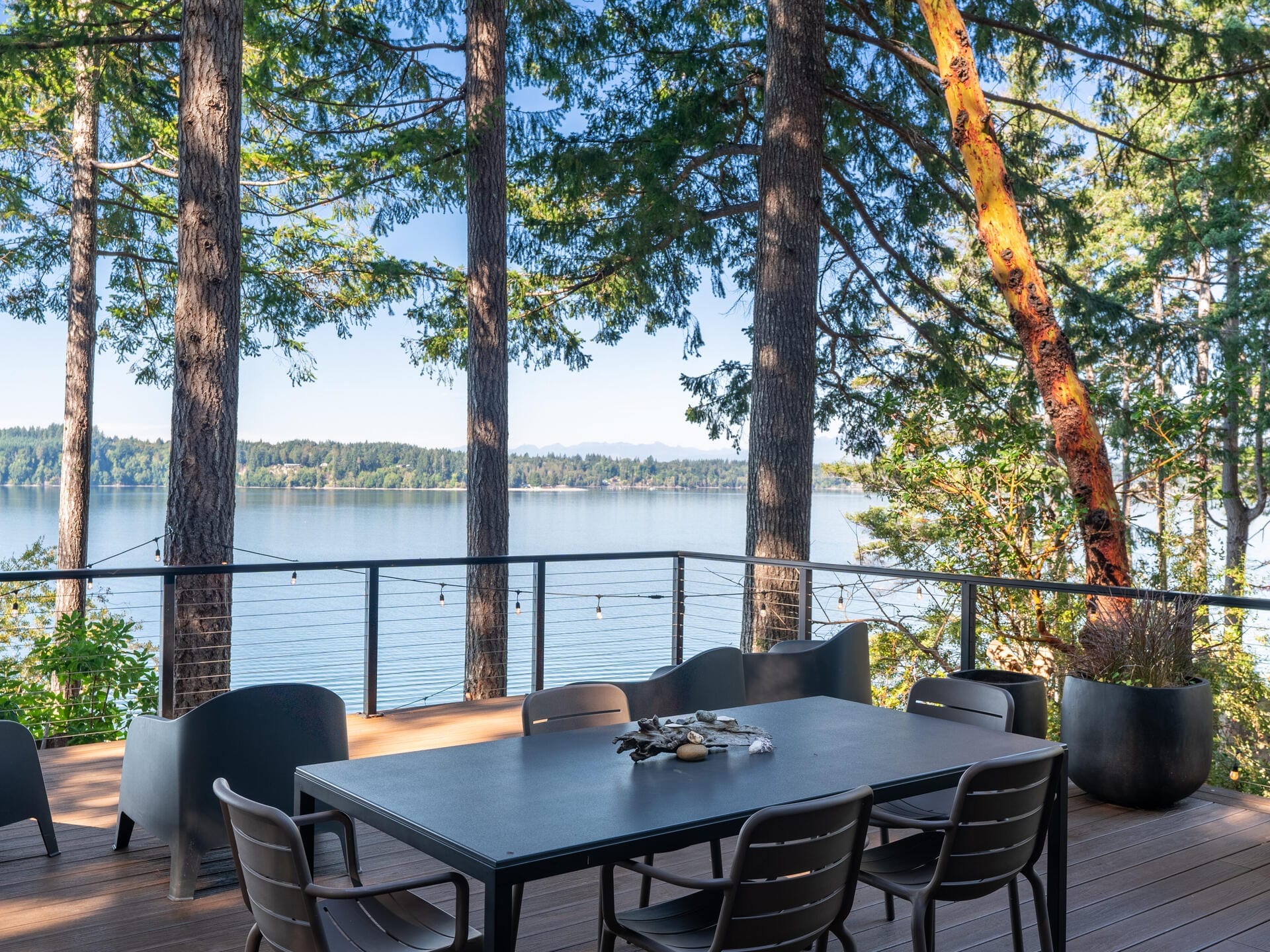 Outdoor patio with a dining table and chairs overlooking a calm lake. Surrounded by tall trees, the scene features a peaceful natural setting with string lights hung along the railing, under a clear blue sky.