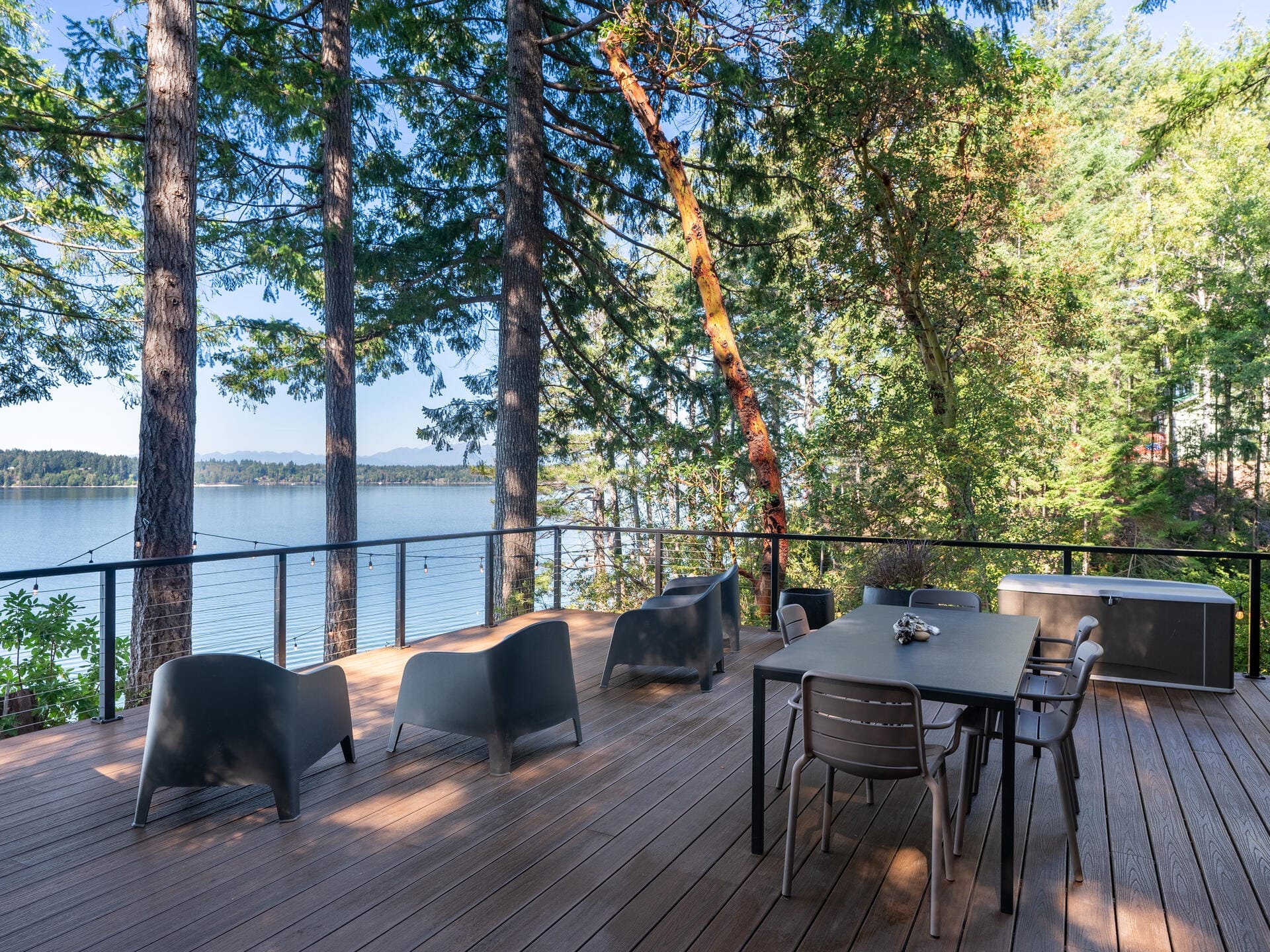 A wooden deck overlooking a tranquil lake surrounded by trees. The deck features modern black chairs, a dining table with chairs, and a bench, creating a serene outdoor space. The lush greenery and calm water enhance the peaceful atmosphere.