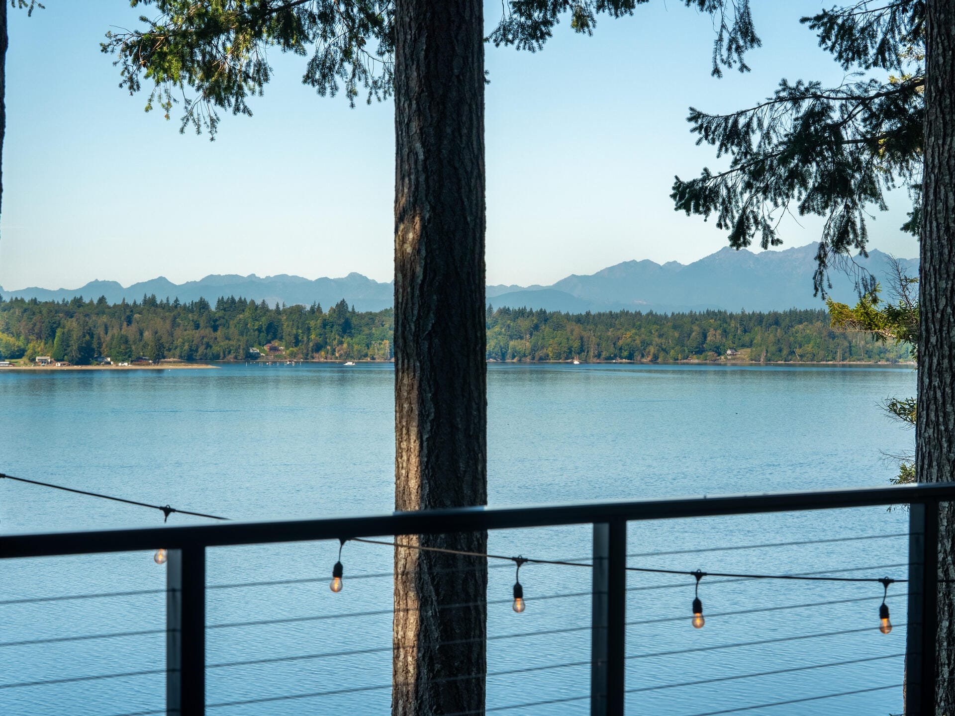 A serene lake view surrounded by trees, with mountains in the distance. The foreground features a railing with string lights, creating a peaceful atmosphere under a clear blue sky.