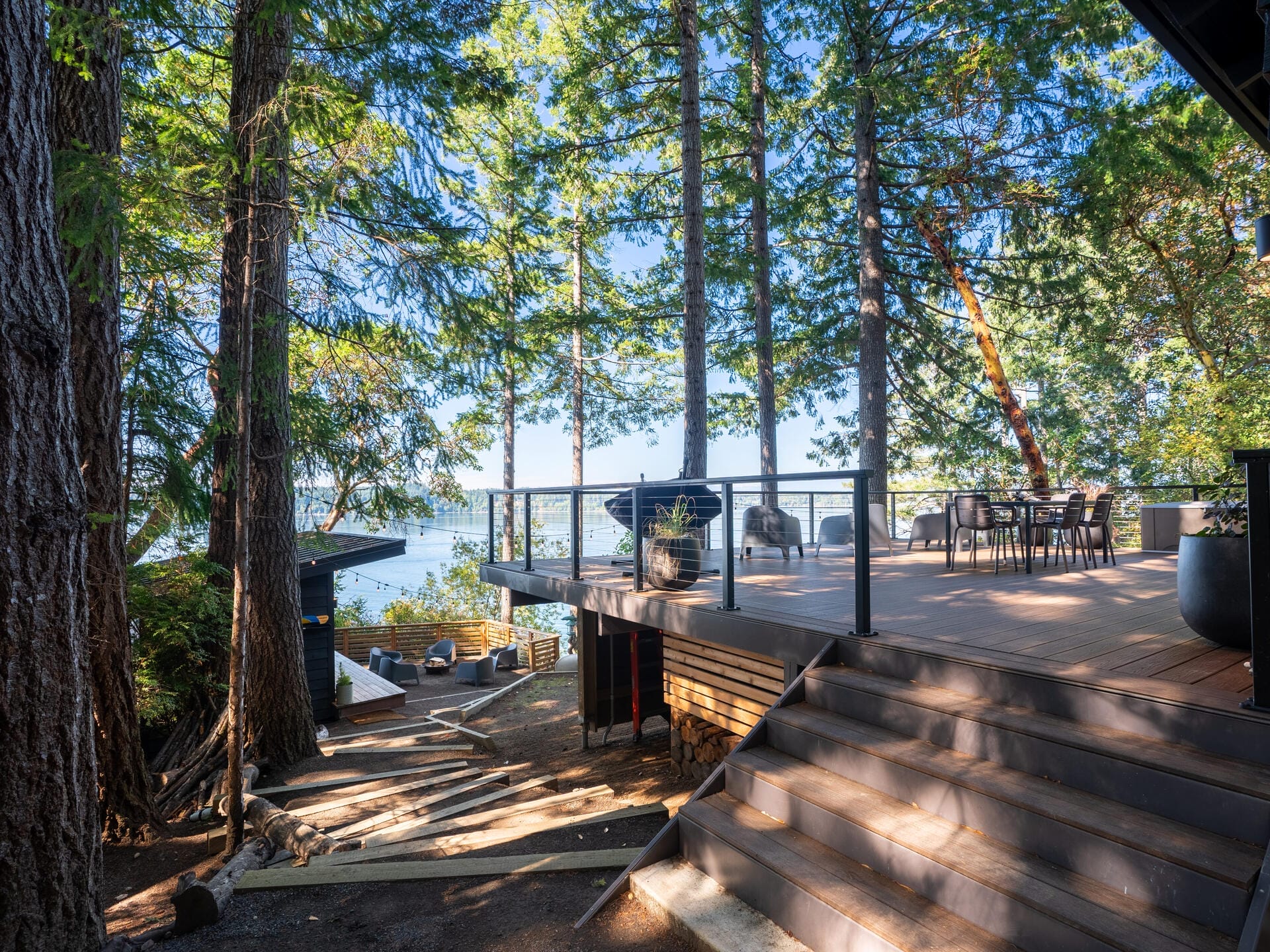 A wooden deck surrounded by tall trees overlooks a peaceful lake. Patio furniture is arranged on the deck, and sunlight filters through the branches, casting dappled shadows. Stairs lead down to a pathway beside the water.