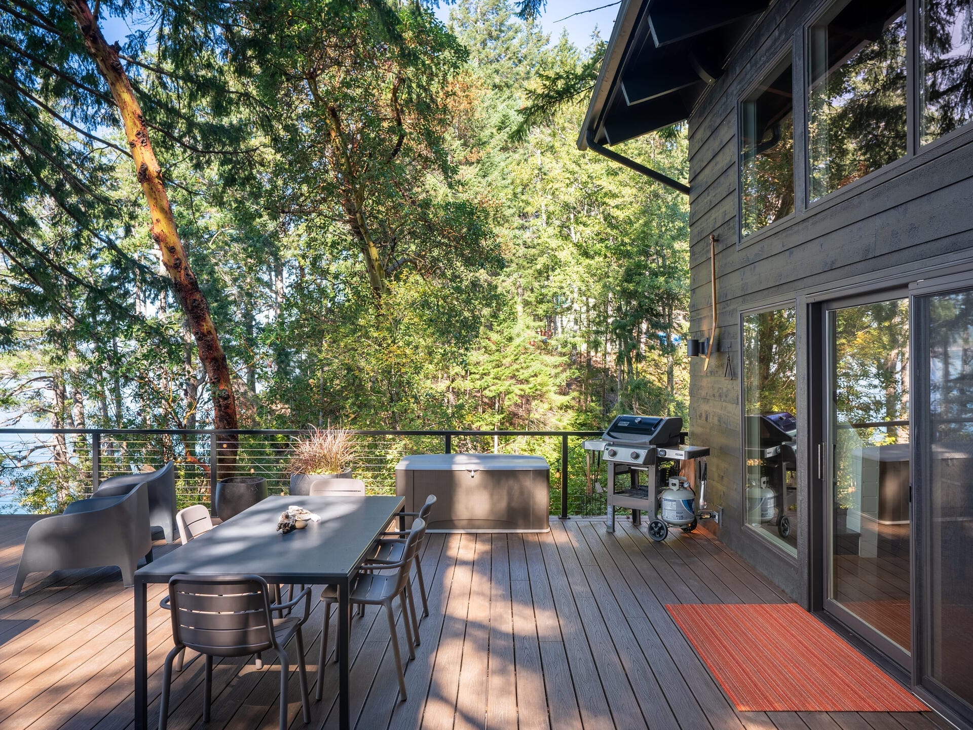 A spacious wooden deck with outdoor furniture, a barbecue grill, and a metal railing overlooks lush green trees. A modern glass door leads inside the house, and a small orange rug is placed near the entrance.