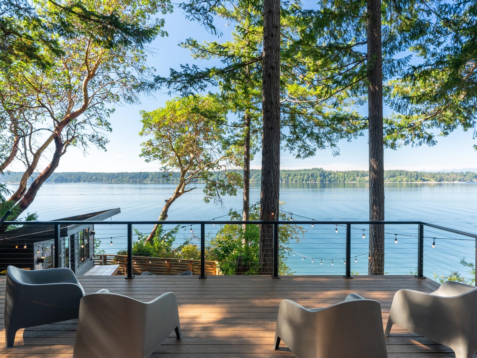 Scenic view of a tranquil lake bordered by dense evergreen trees. An outdoor deck features modern seating and is lined with a black railing. String lights hang nearby, and the clear sky reflects on the calm water.