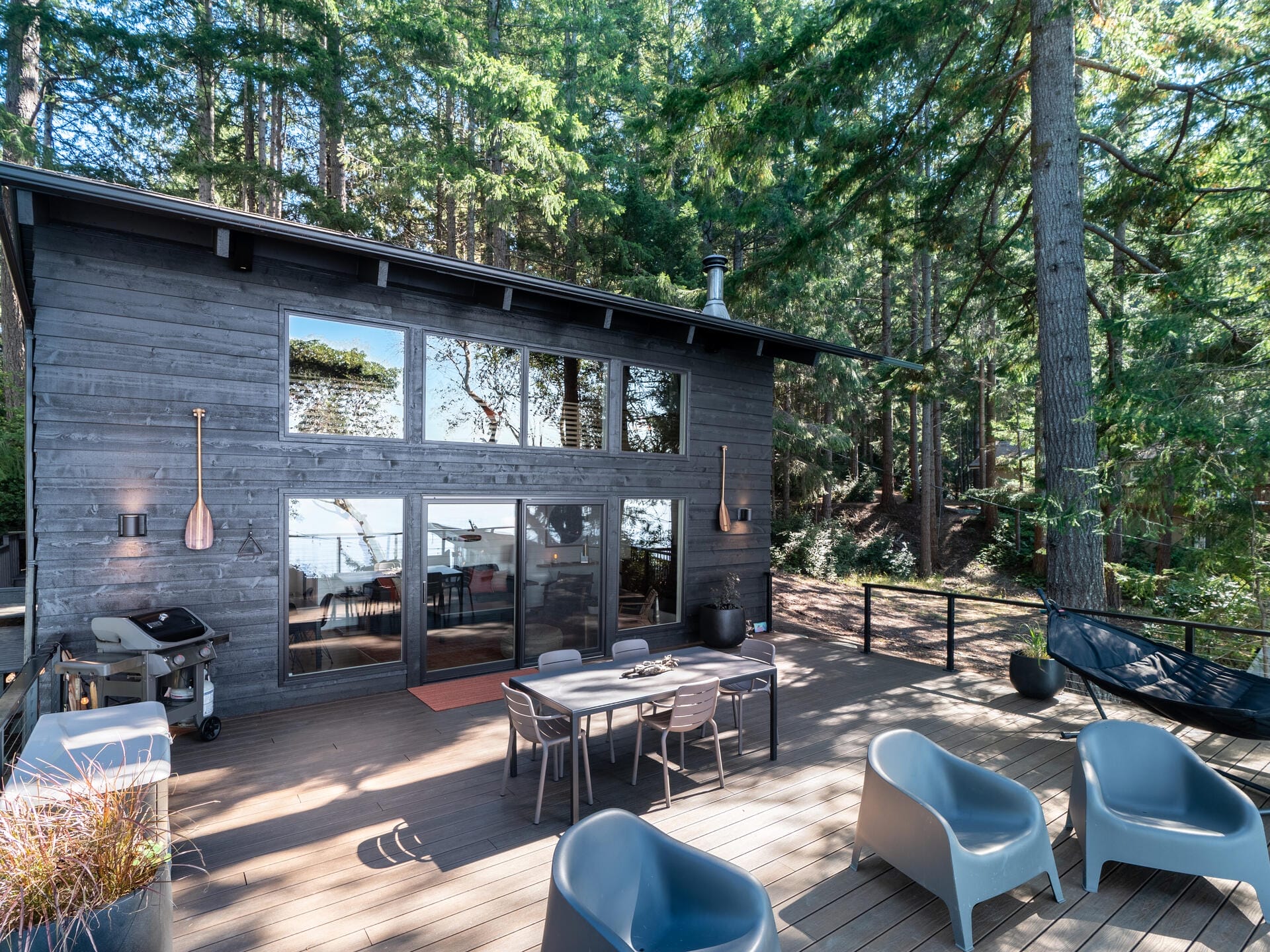 A modern cabin with a wooden deck in a forest setting. The deck features outdoor seating, a grill, and a table with chairs. The cabin is dark wood with large windows reflecting trees. Tall evergreens surround the area, creating a serene atmosphere.