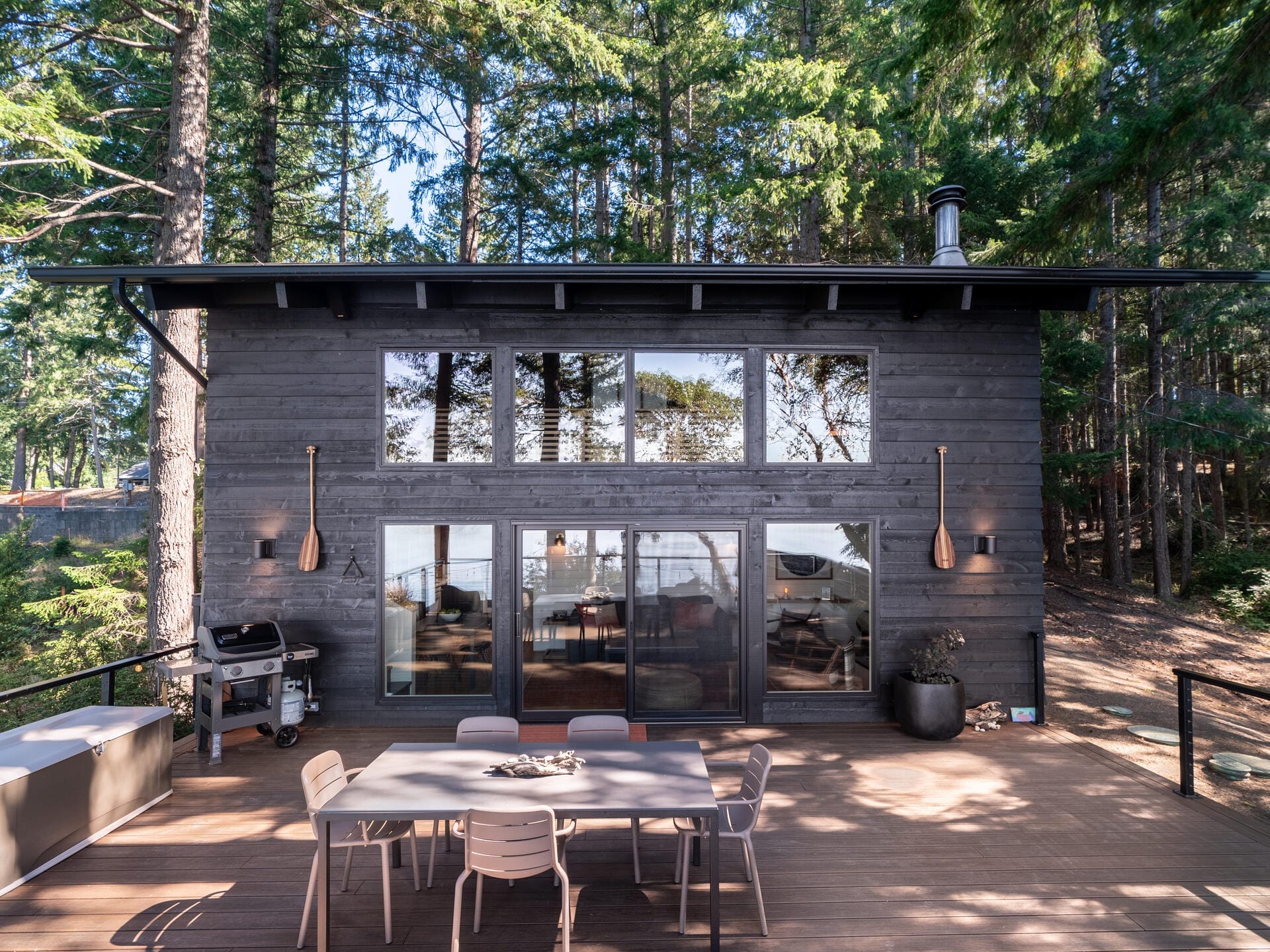 A modern black wooden cabin with large windows in a forest setting. The front patio features a dining table, chairs, and a grill. Tall trees surround the cabin, and two wooden paddles are mounted as decor.