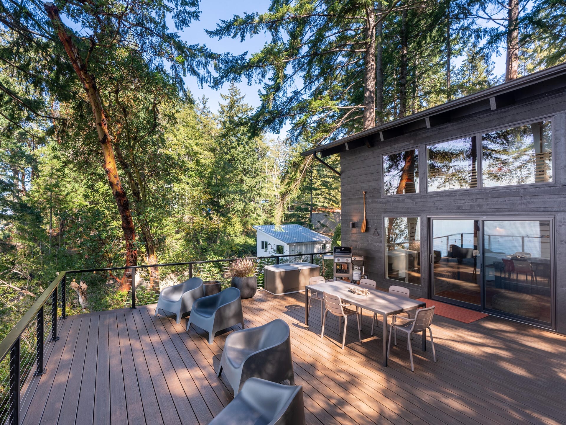 A spacious wooden deck surrounded by tall trees features modern gray chairs and a dining table. A contemporary house with large windows is on the right, and a view of distant water is visible through the trees.