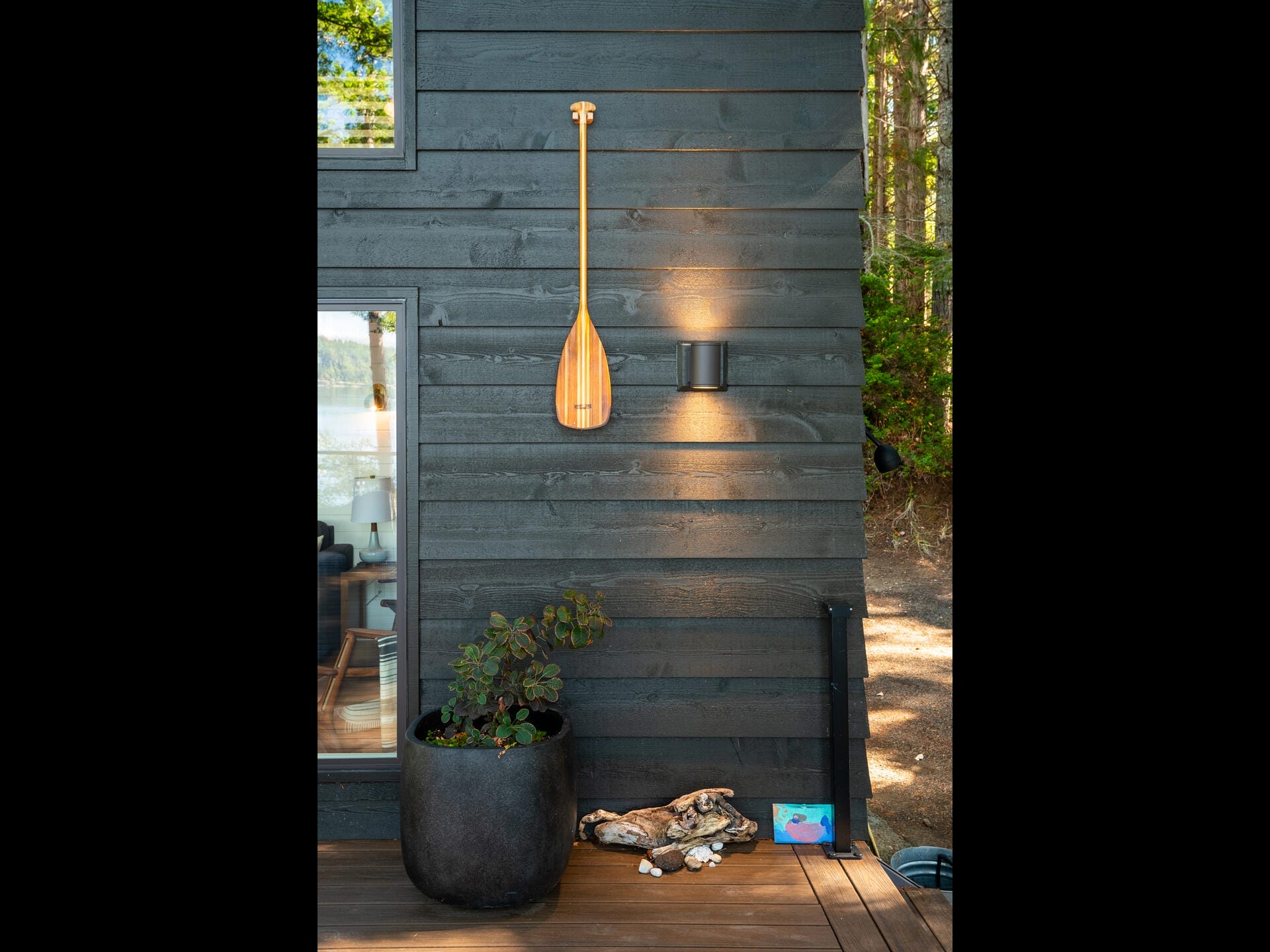 A wooden paddle decoratively hangs on the dark exterior wall of a house. Below, a black outdoor light fixture illuminates a plant in a large pot. Various small items are placed on the wooden deck, including toys. Trees are visible in the background.