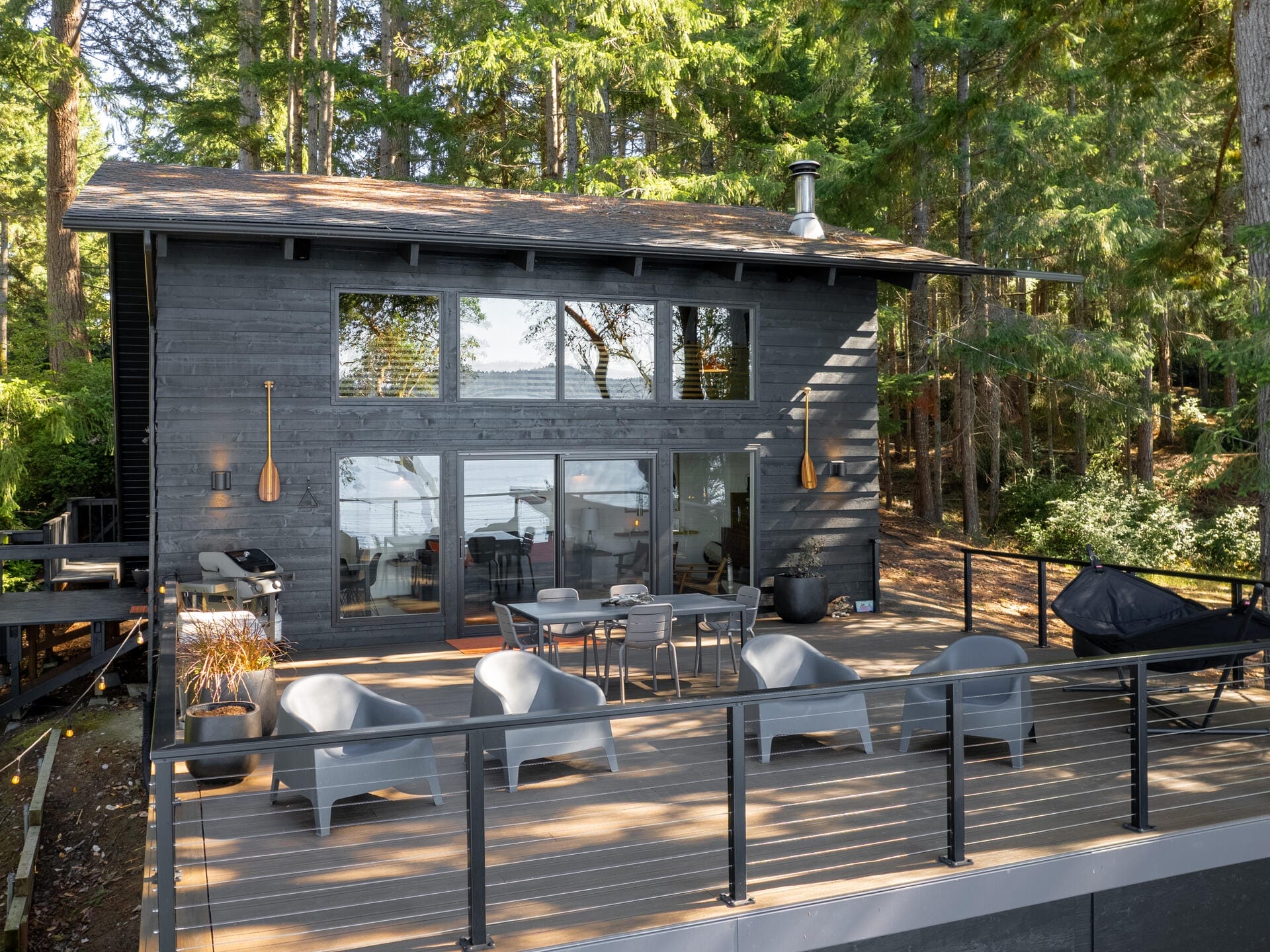 A modern cabin with large windows nestled in a forested area. It features a spacious deck with gray chairs and a table, overlooking a serene lake. Tall trees surround the house, adding to the tranquil atmosphere.