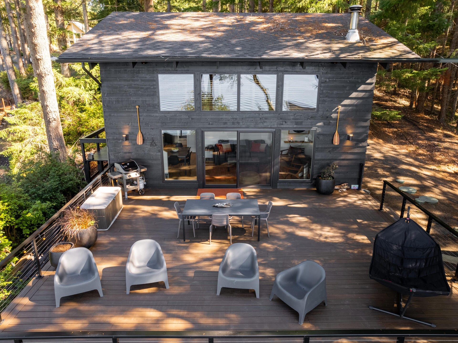 A modern two-story house with large windows, set in a forested area. A spacious wooden deck features a dining table with chairs, four gray lounge chairs, a barbecue grill, and a hammock swing. Trees surround the house, creating a serene atmosphere.