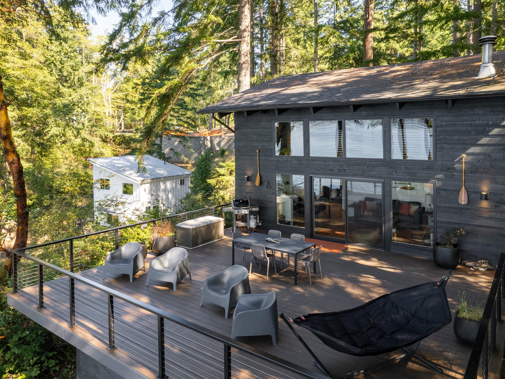 A modern cabin with a spacious wooden deck nestled among tall trees. The deck features grey outdoor chairs, a table, a grill, and a hammock. Large windows reflect the surrounding forest. Another house is visible in the background.