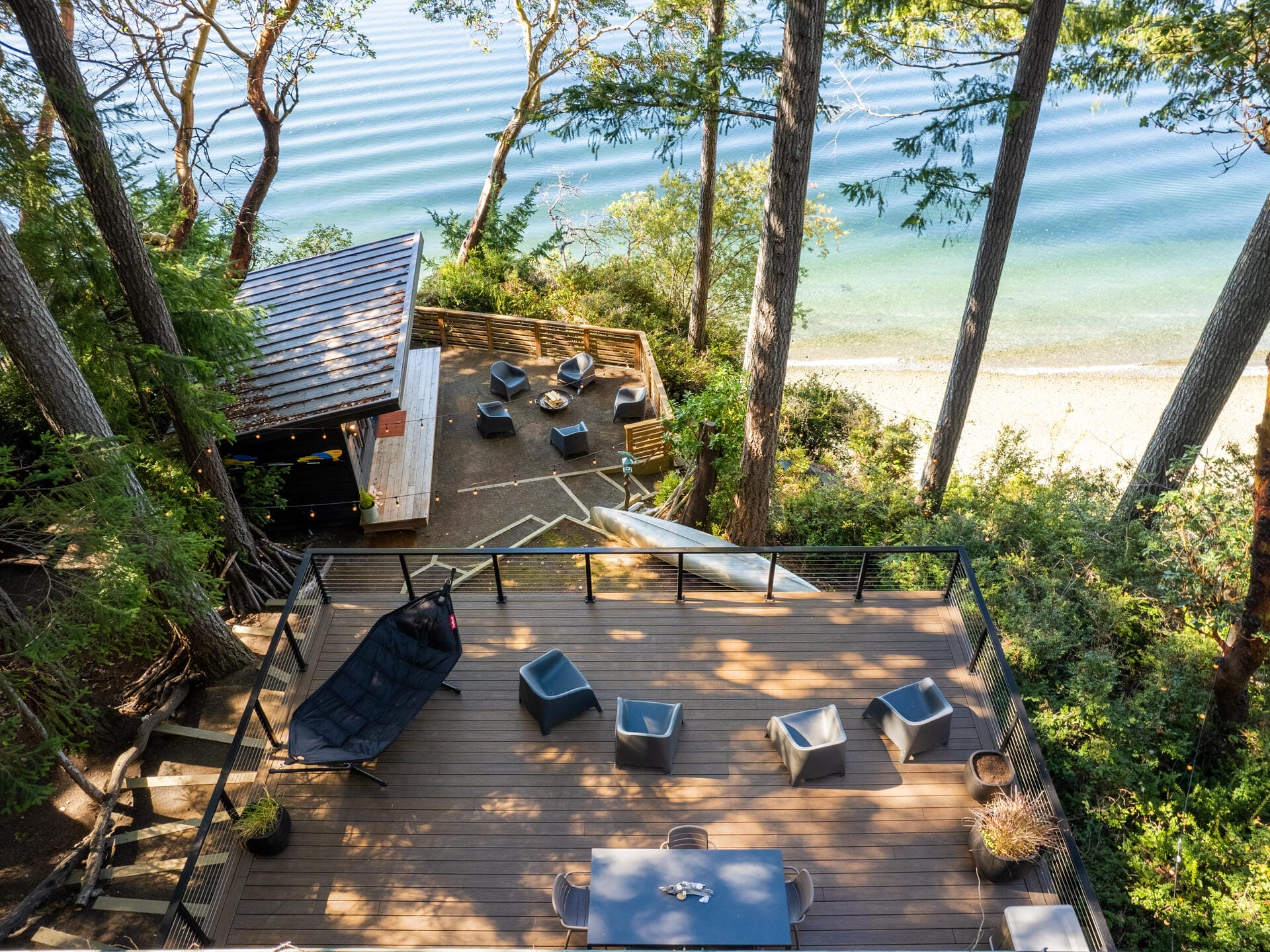 Aerial view of a wooden deck with modern chairs overlooking a serene beach and calm water. Surrounded by tall trees, theres an outdoor seating area below with more chairs and a small shelter, creating a picturesque and relaxing coastal retreat.