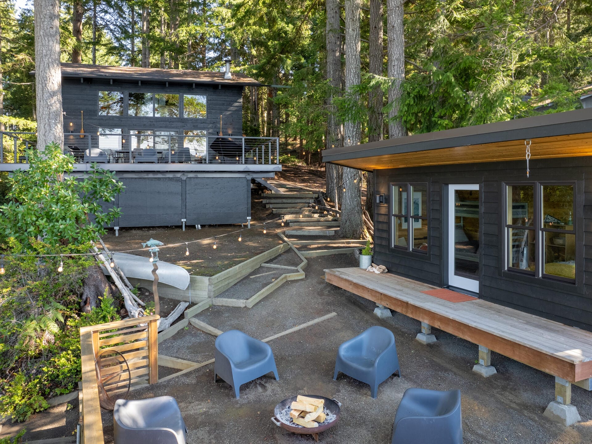 A cozy outdoor area with a fire pit surrounded by four blue chairs. Two modern black wooden cabins are nestled among tall trees. One cabin has a large deck and glass doors. The setting is serene and surrounded by lush greenery.