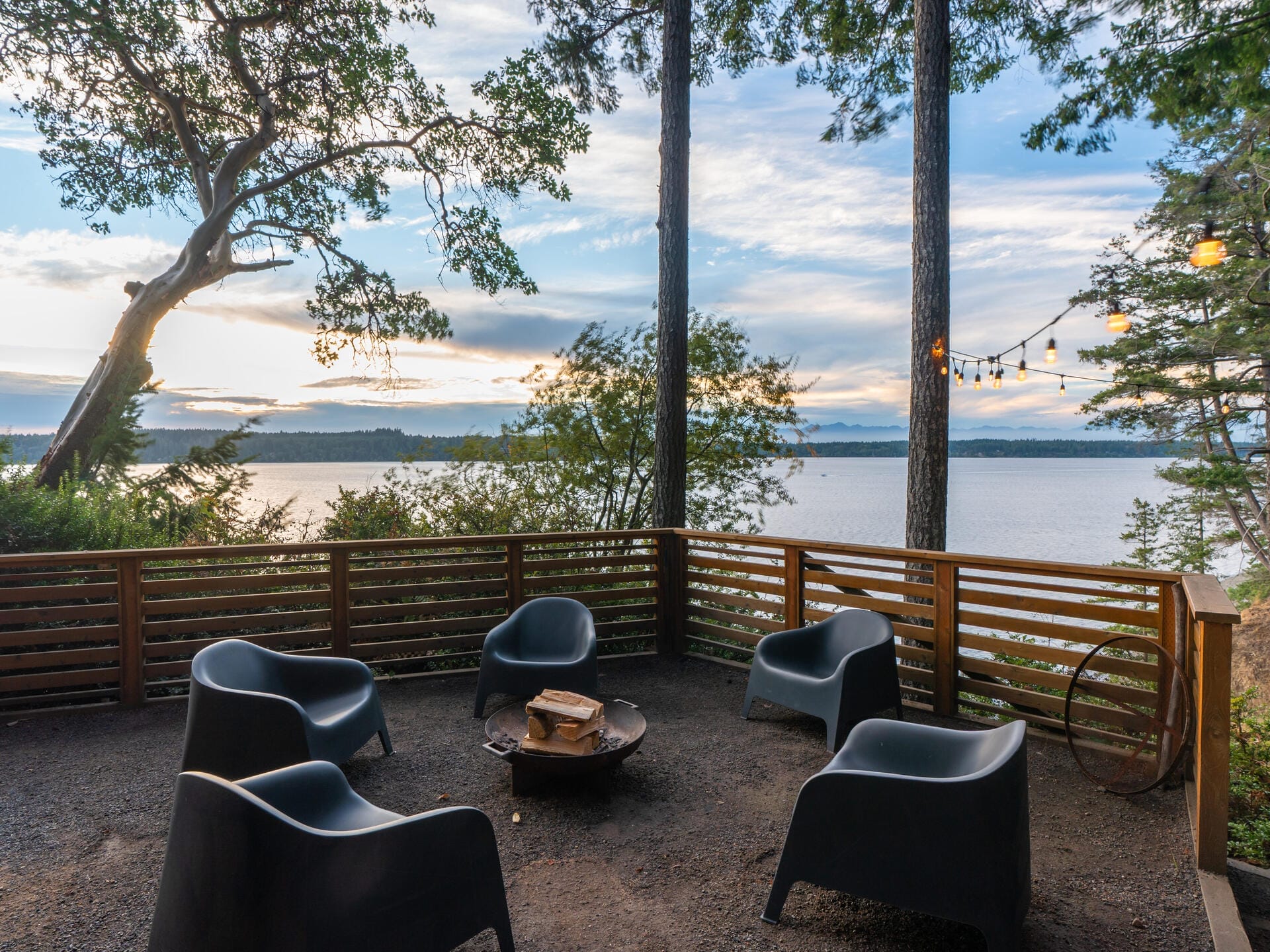 A scenic outdoor deck overlooking a lake, surrounded by trees. Four modern black chairs are arranged around a circular fire pit. String lights hang above, creating a cozy atmosphere under a cloudy sky at sunset.