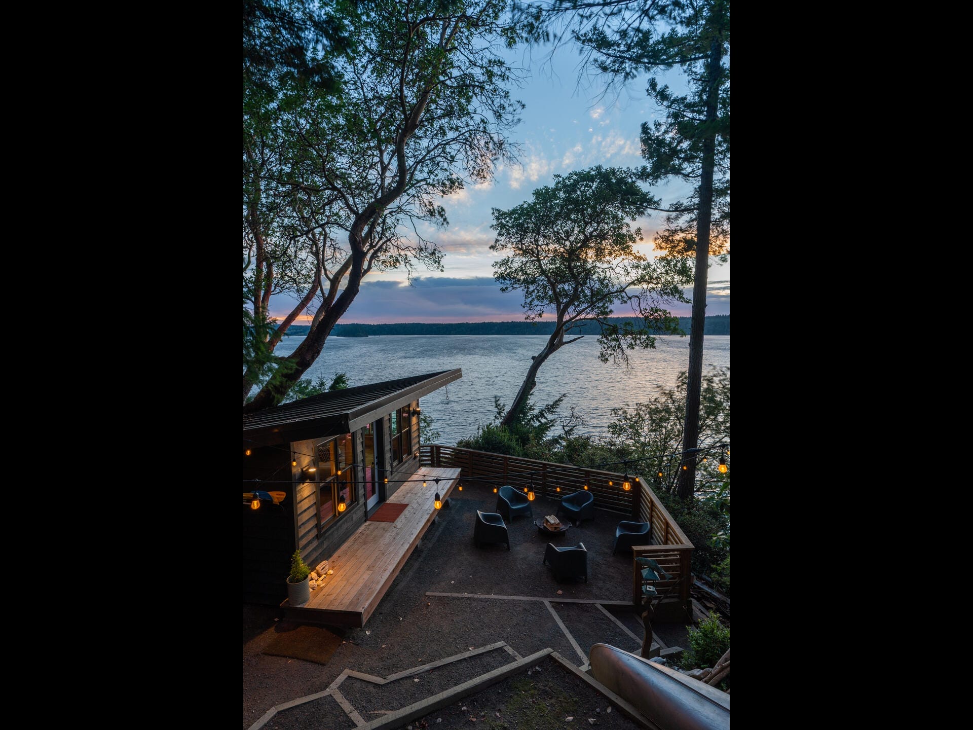 A cozy lakeside cabin with a deck overlooking the water at sunset. The deck features outdoor seating and ambient lighting, surrounded by trees. The sky is painted with hues of orange and blue as the sun sets over the horizon.