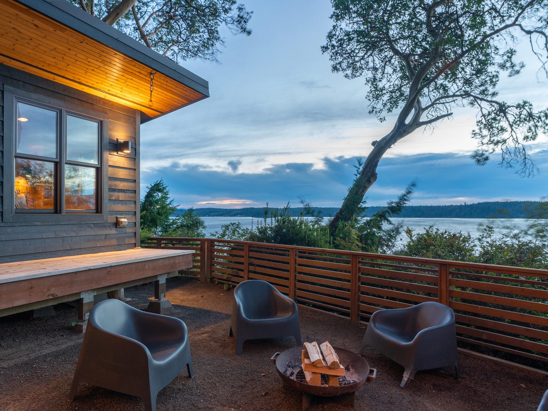 Cozy outdoor seating area with four modern chairs around a fire pit, beside a wooden deck. Overlooks a serene body of water with distant mountains and a dramatic, cloudy sky during sunset. Trees frame the view, adding to the tranquil atmosphere.