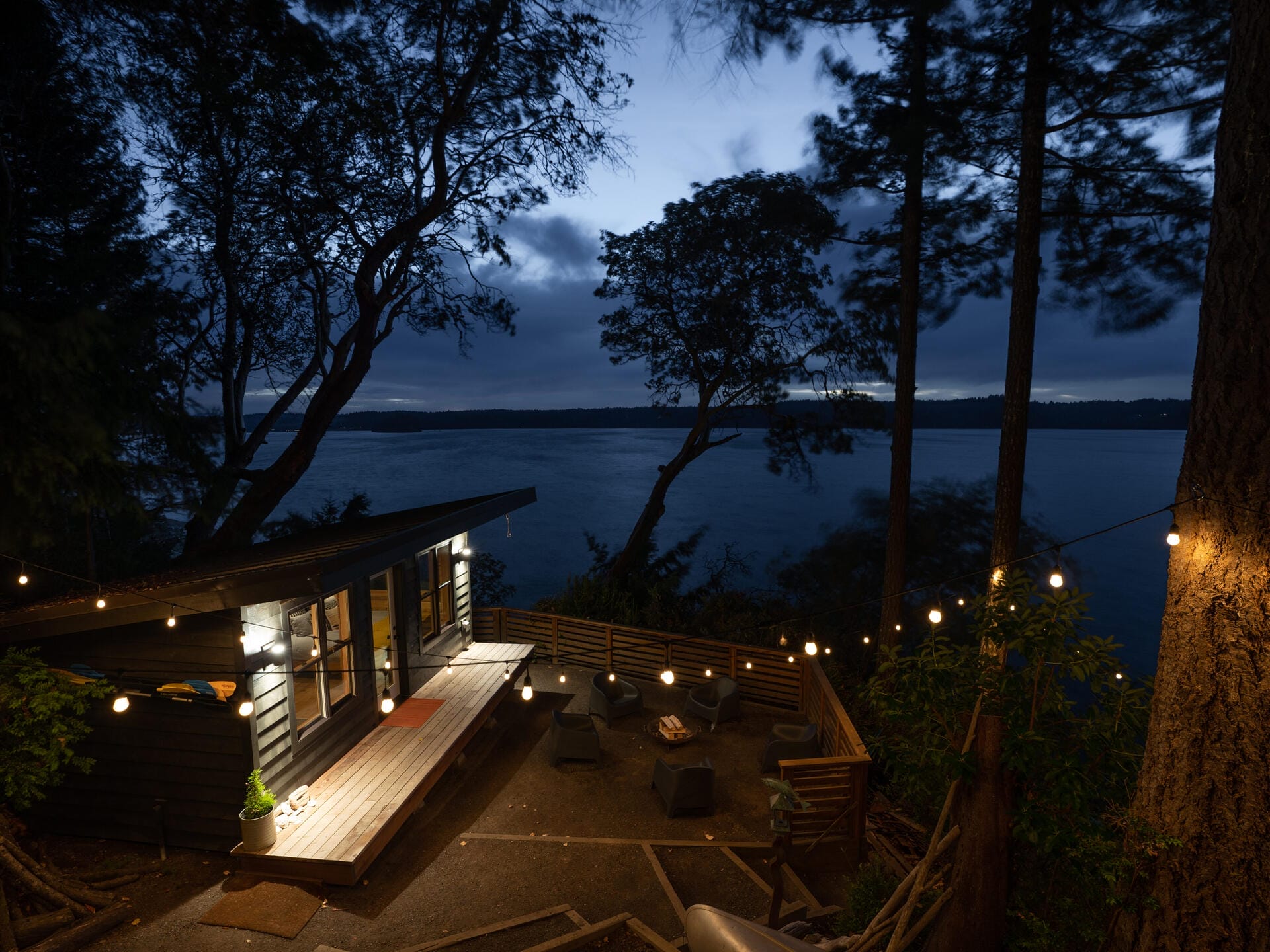 A cozy cabin surrounded by trees at dusk, overlooking a calm body of water. The warm glow from the cabin windows and string lights creates a serene atmosphere. Outdoor seating is arranged on a deck beneath the lights.