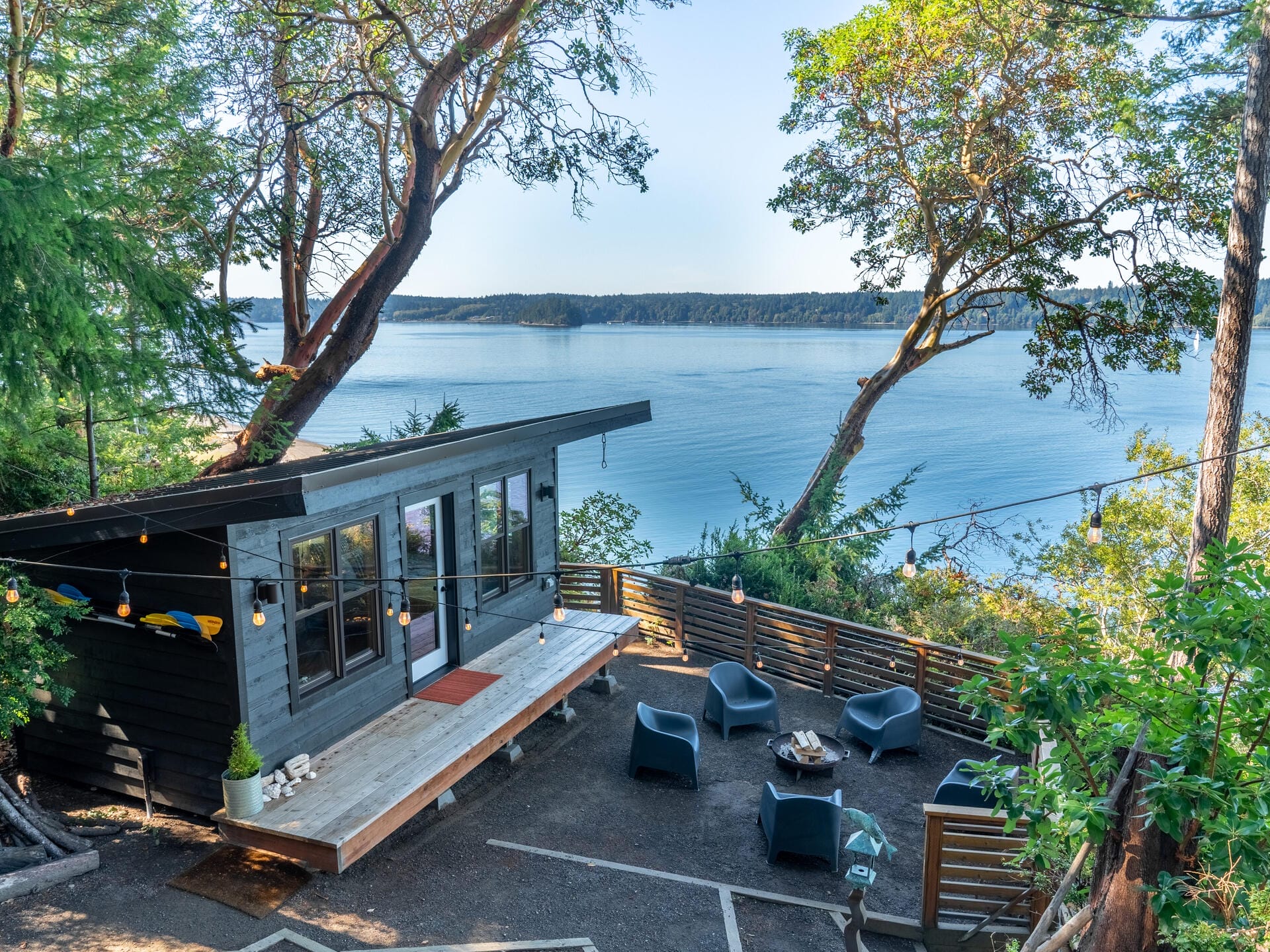 A modern cabin with large windows overlooks a serene body of water surrounded by trees. The outdoor deck features seating and a fire pit. String lights are strung above the patio, creating a cozy atmosphere with a backdrop of distant shoreline.