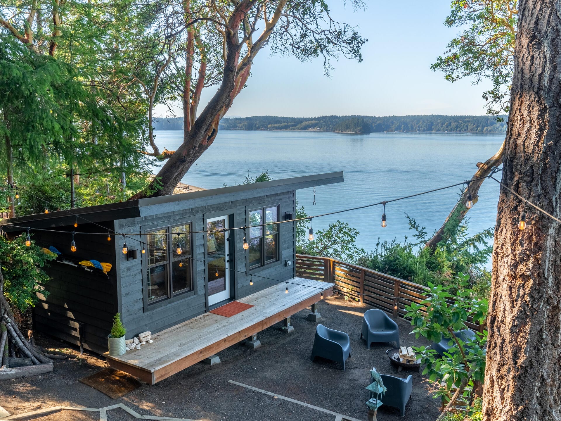 A modern cabin with large windows is surrounded by trees, overlooking a peaceful body of water. A patio with string lights, chairs, and a table sits nearby, creating a serene outdoor space.