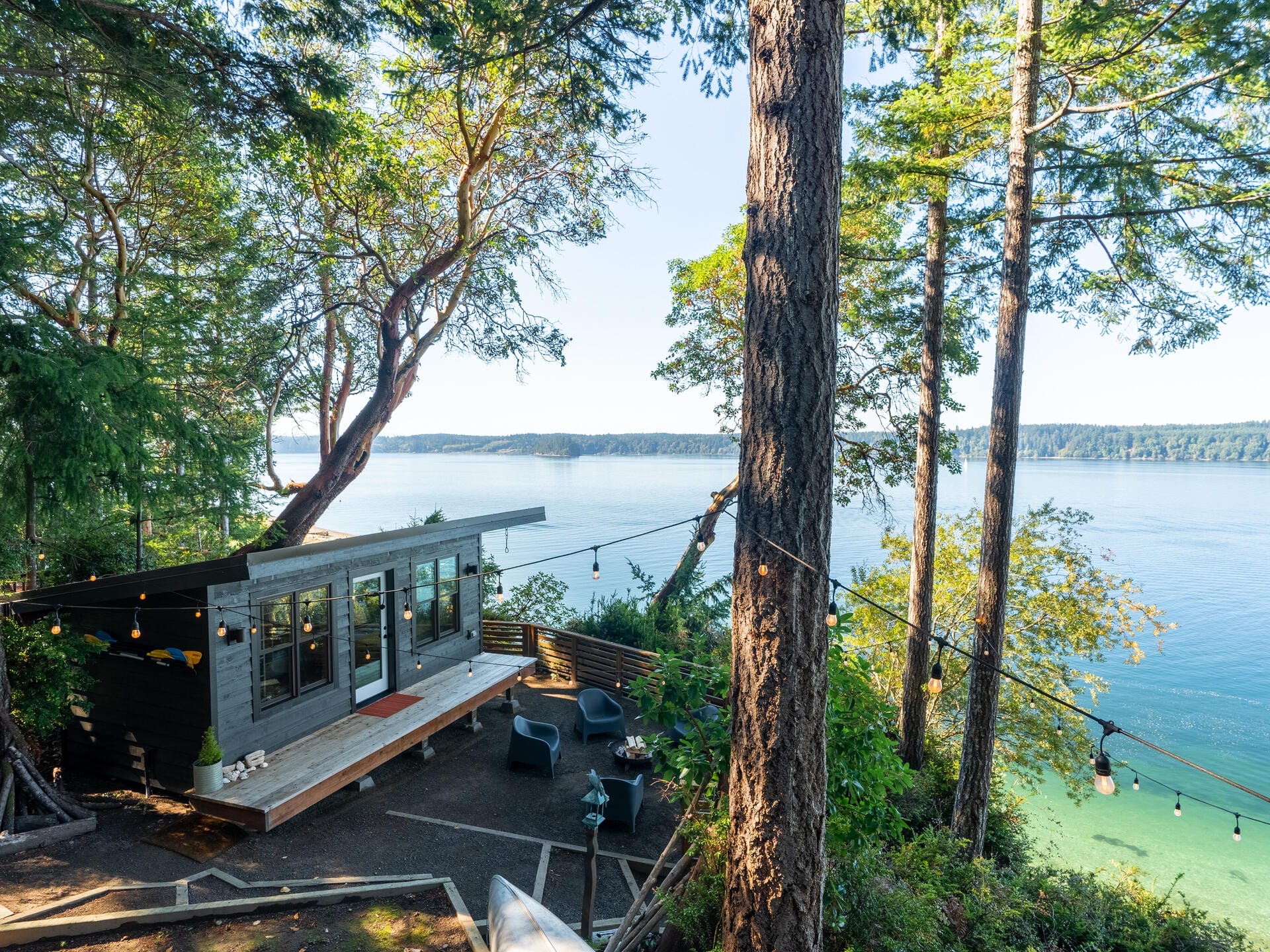 A modern cabin sits on a hillside surrounded by tall trees, overlooking a body of water. The patio features outdoor seating and string lights, creating a serene atmosphere. The sunlit scene offers a picturesque view of the landscape.