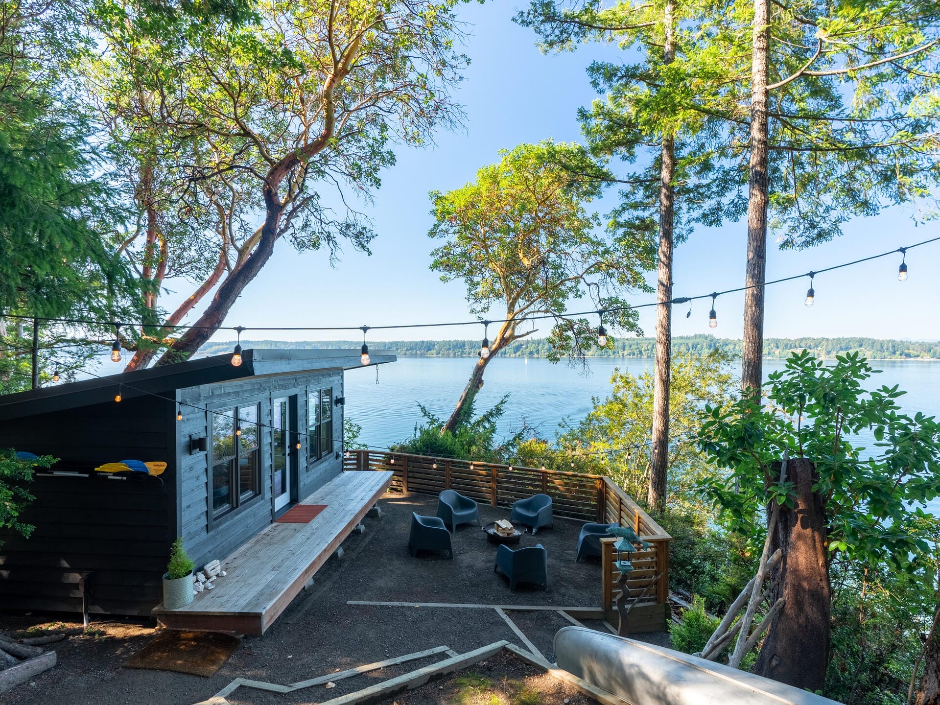 A modern, dark-paneled cabin with large windows sits near a scenic lake surrounded by lush trees. Outdoor seating and string lights enhance the tranquil setting. The view showcases the calm water and distant shore under a clear blue sky.