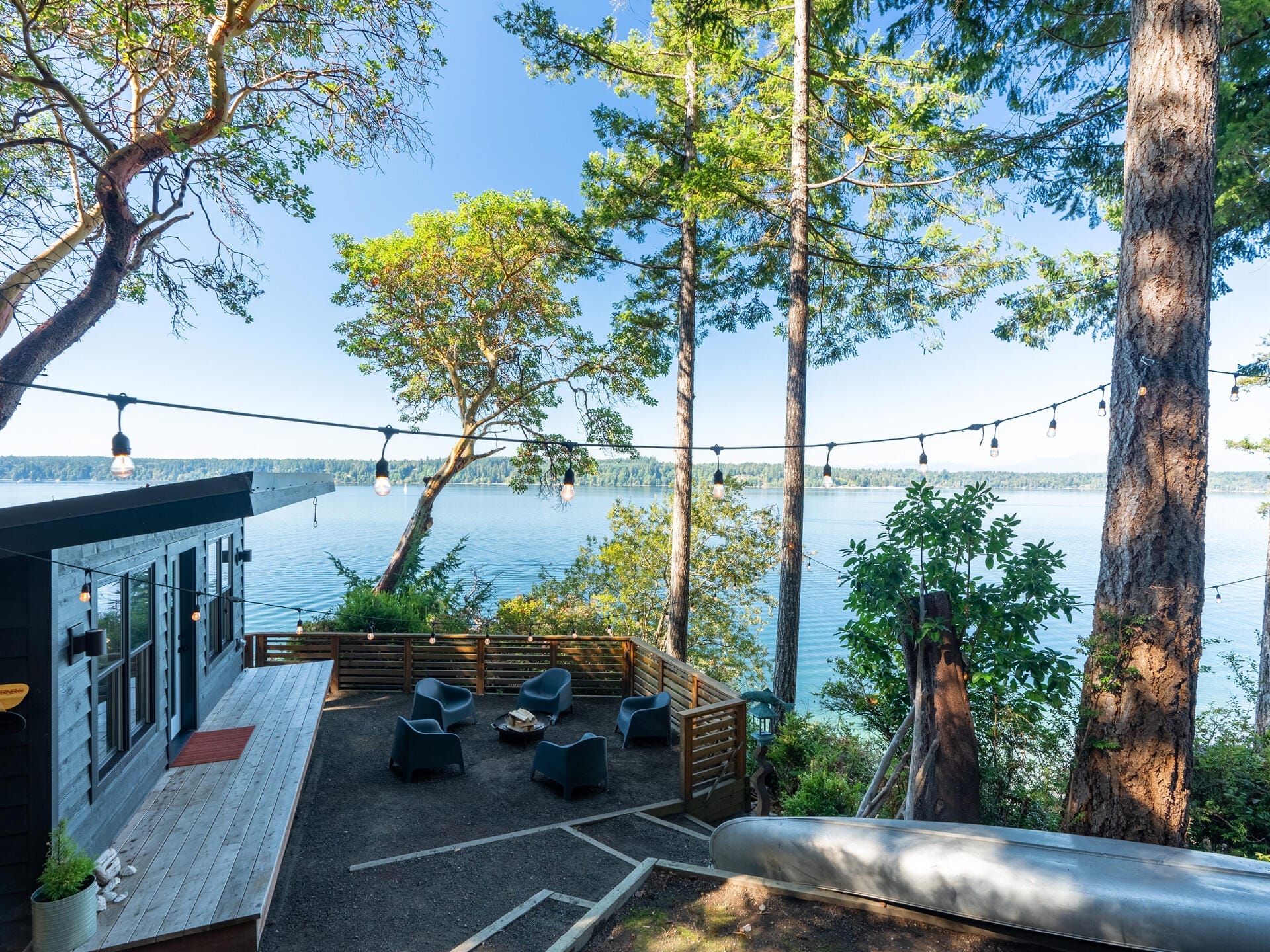 A scenic view of a lakeside patio surrounded by tall trees and overlooking a serene body of water. The patio features several chairs around a fire pit, string lights, and a small building with large windows. A canoe rests nearby.