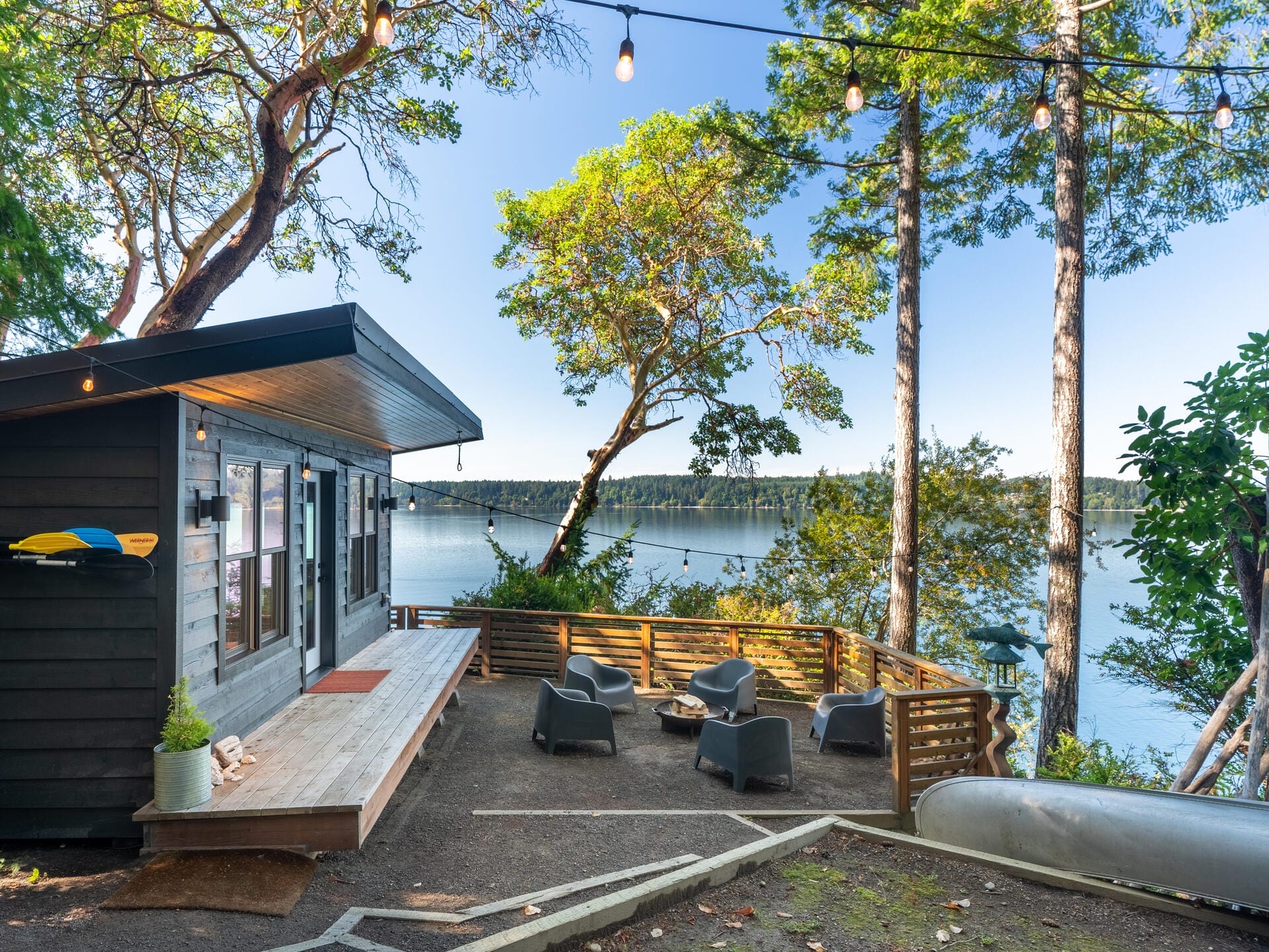 A scenic patio overlooks a tranquil lake surrounded by trees. The area features outdoor seating, string lights, and a canoe. A modern, dark-colored building stands to the left. The setting suggests a peaceful, natural retreat.