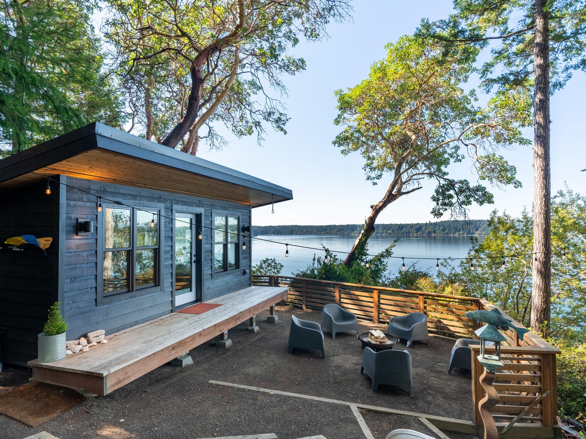 Modern cabin with large windows overlooking a serene lake. Surrounded by tall trees, the area features a wooden deck with gray lounge chairs and a small fire pit, offering a peaceful, natural setting.