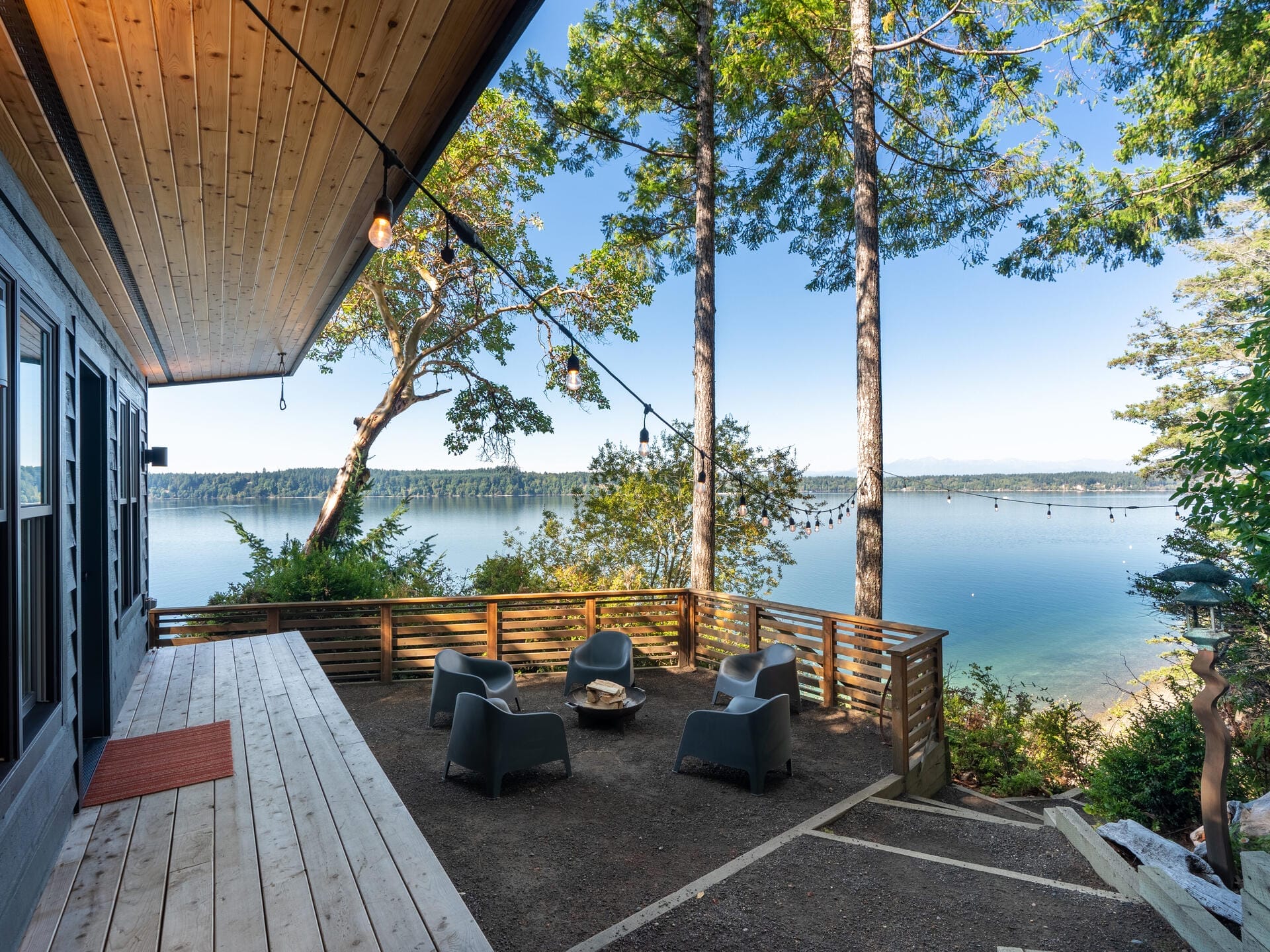 Scenic lakeside view from a wooden deck with outdoor seating. The area is surrounded by tall trees and overlooks a calm body of water under a clear blue sky. Hanging lights add a cozy ambiance to the peaceful setting.