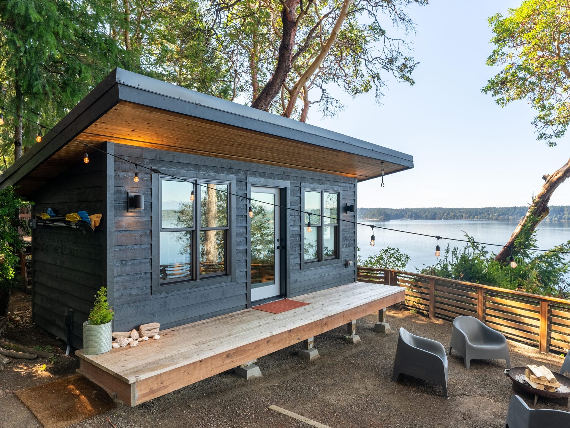 A modern, small cabin with large windows on a wooded lakeside. The cabin is dark-colored with a flat roof, surrounded by trees. String lights hang above a small deck, where two chairs and a table are placed. The lake is visible in the background.