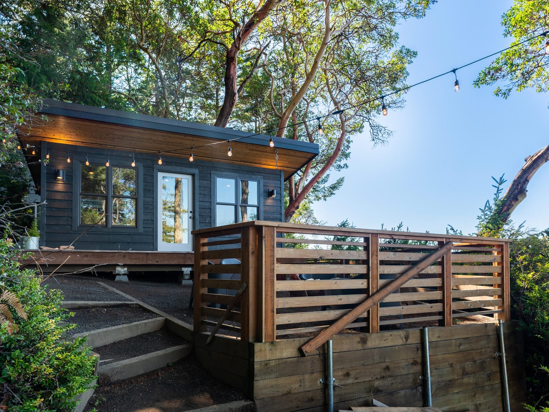 A modern cabin with dark wood exterior, large glass doors, and windows is nestled among trees. String lights hang above a wooden deck with a railing, and steps lead up to the entrance. The sky is clear, creating a serene atmosphere.