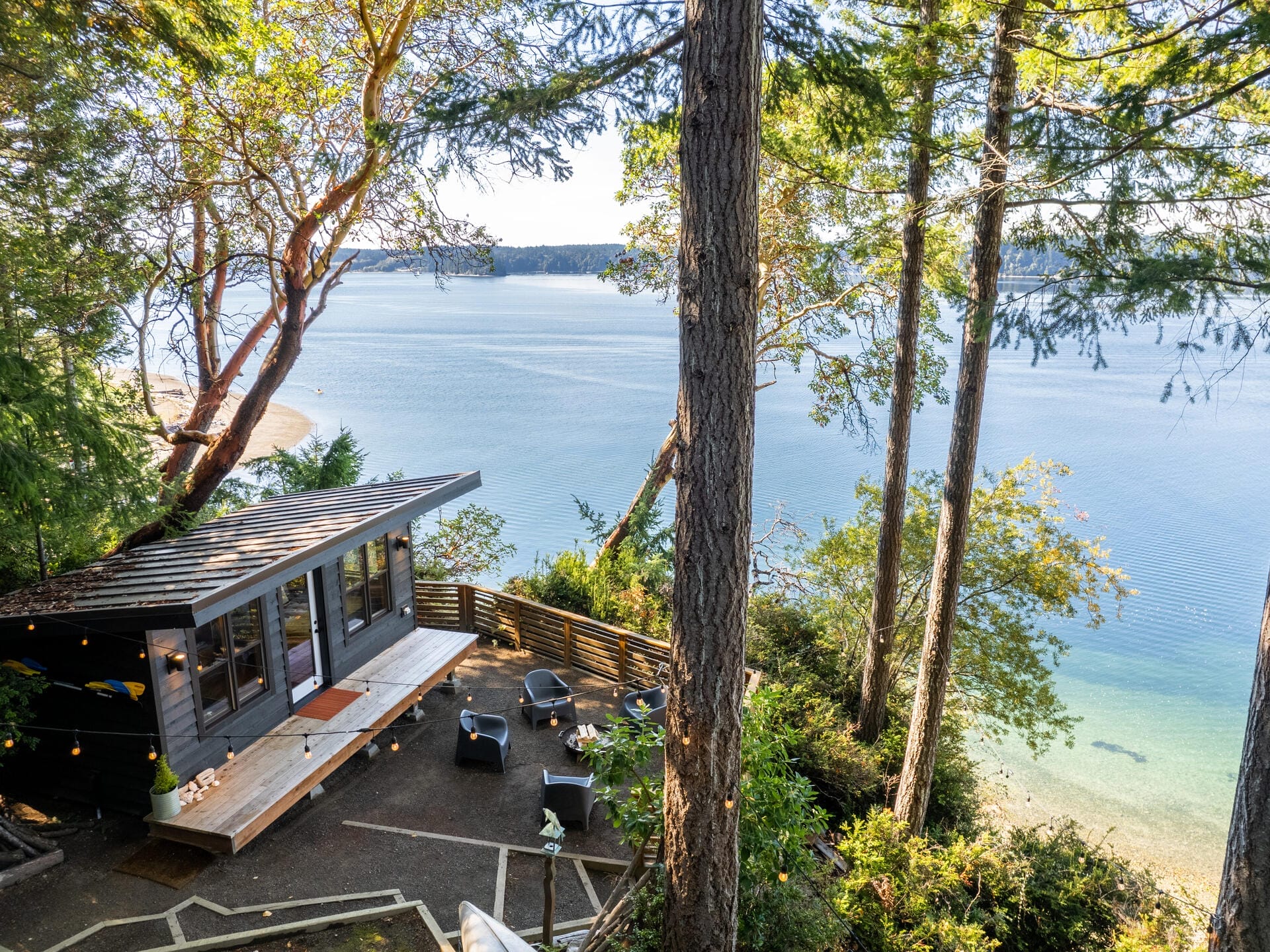 A modern, small cabin with large windows is nestled among tall trees overlooking a serene body of water. The patio area has seating and string lights, surrounded by lush greenery. The sky is clear, and the water reflects the sunlight.