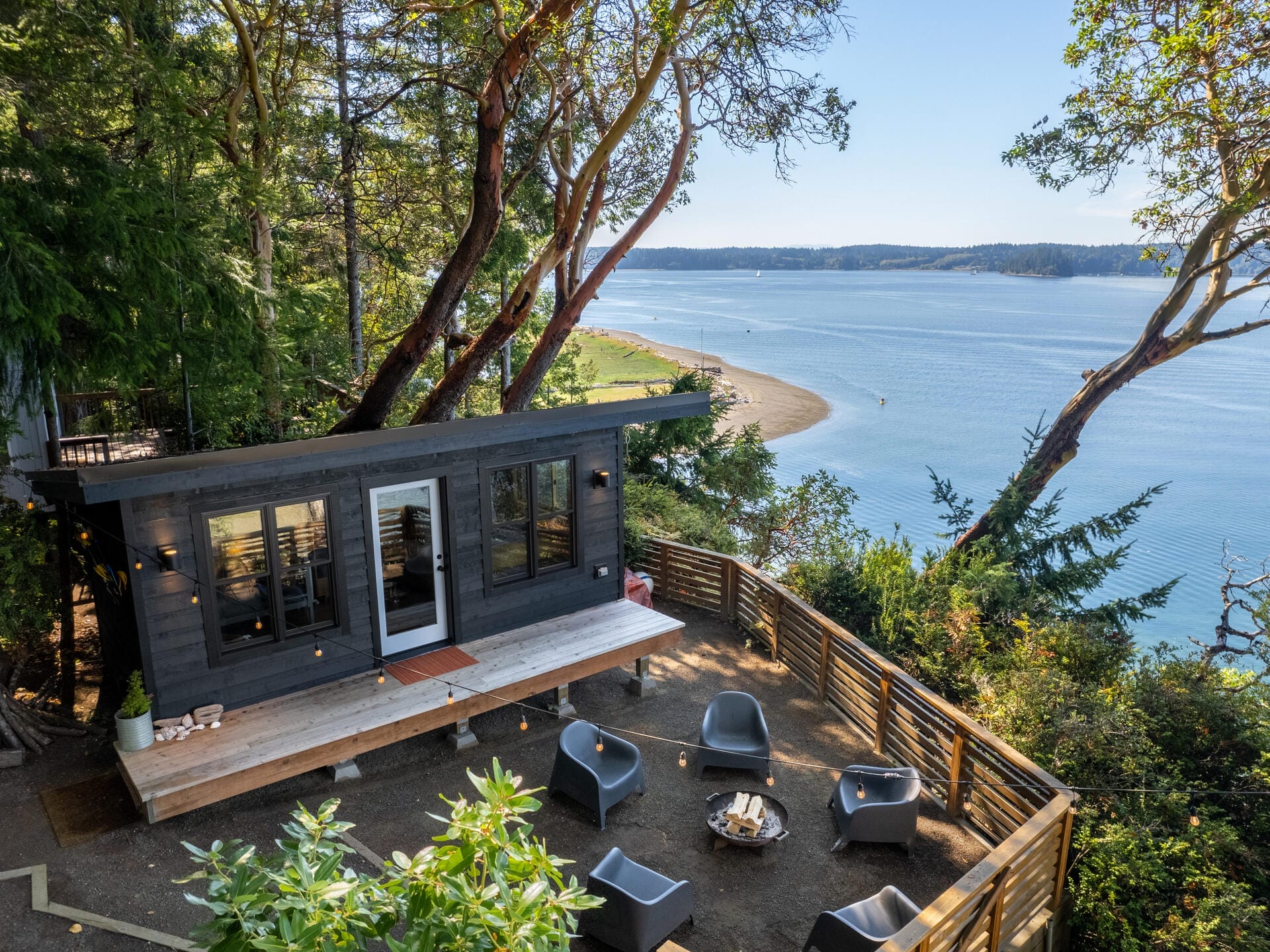 A modern cabin with a large deck is nestled among trees overlooking a serene, blue body of water. The outdoor seating area has several chairs around a fire pit. A railing lines the deck, offering stunning views of the landscape.