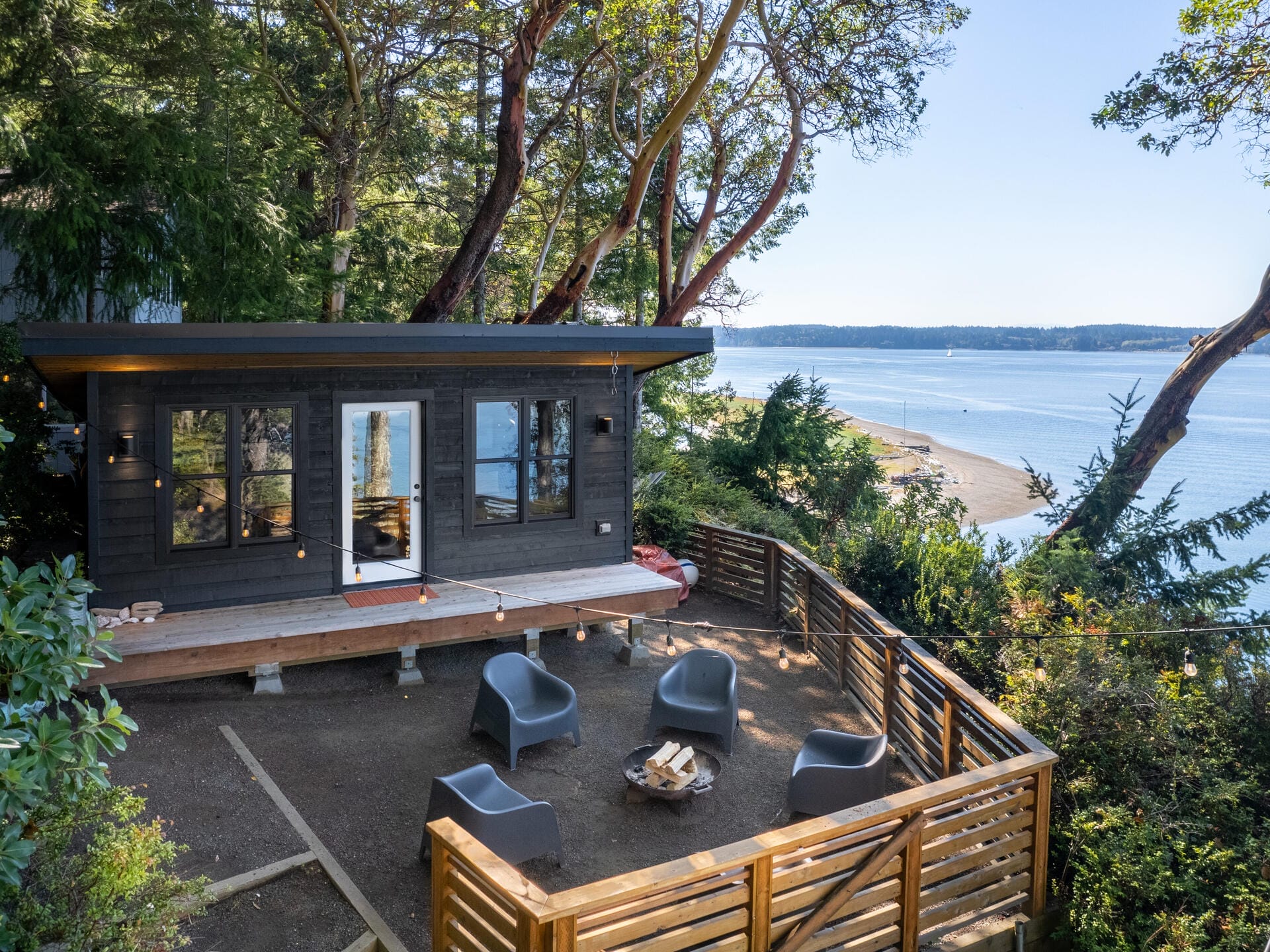 A small, modern cabin with a wooden deck is nestled among trees overlooking a tranquil body of water. The outdoor area features gray chairs and a fire pit, enclosed by a wooden fence, providing a peaceful, natural retreat.