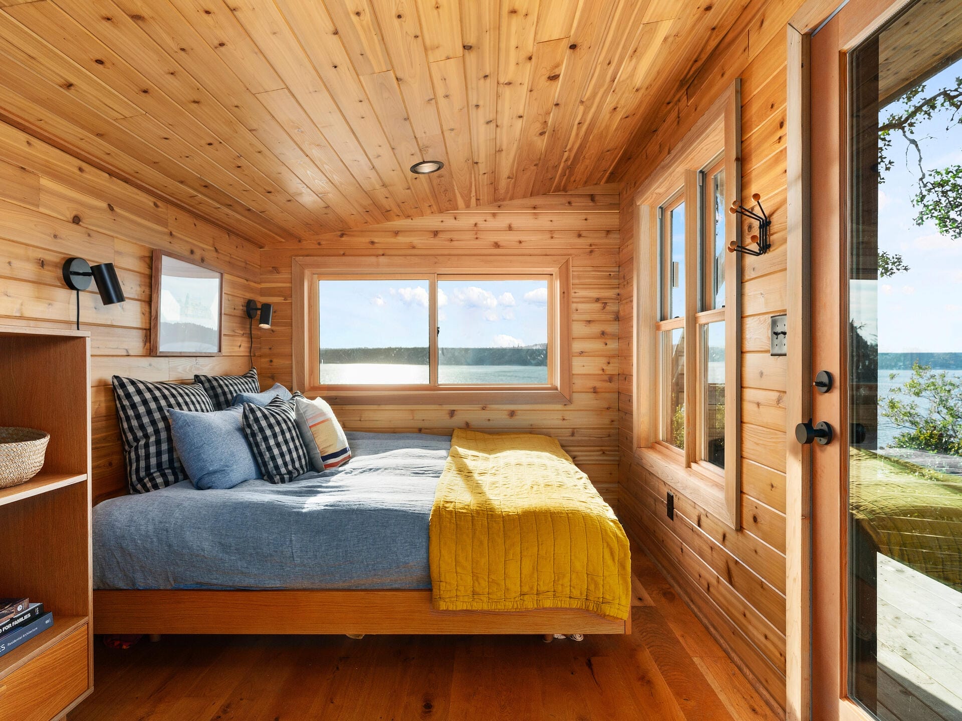 A cozy wooden cabin bedroom with a large window providing a view of a sunny lake. The room features a neatly made bed with a blue comforter and a yellow throw. Bright natural light fills the room, highlighting the warm wood paneling.